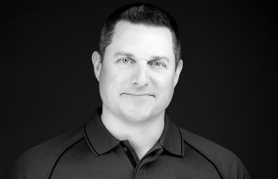 Black and white portrait of a man with short dark hair, wearing a collared shirt, smiling slightly against a dark background.