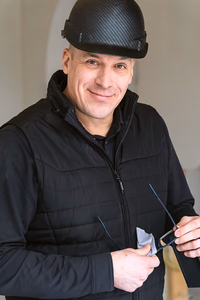 Smiling man wearing a black quilted vest and carbon fiber textured black safety helmet, holding glasses and a cleaning cloth in an indoor setting.