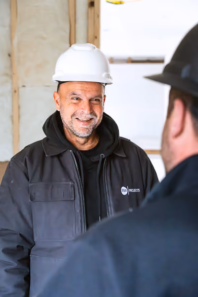 Smiling construction worker wearing a white hard hat and dark jacket with a logo, engaged in conversation with a colleague in a black hard hat inside a building under construction.
