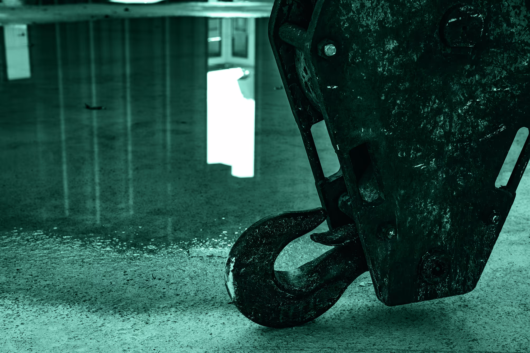 Close-up of a large, weathered metal hook hanging above a polished concrete floor reflecting light.