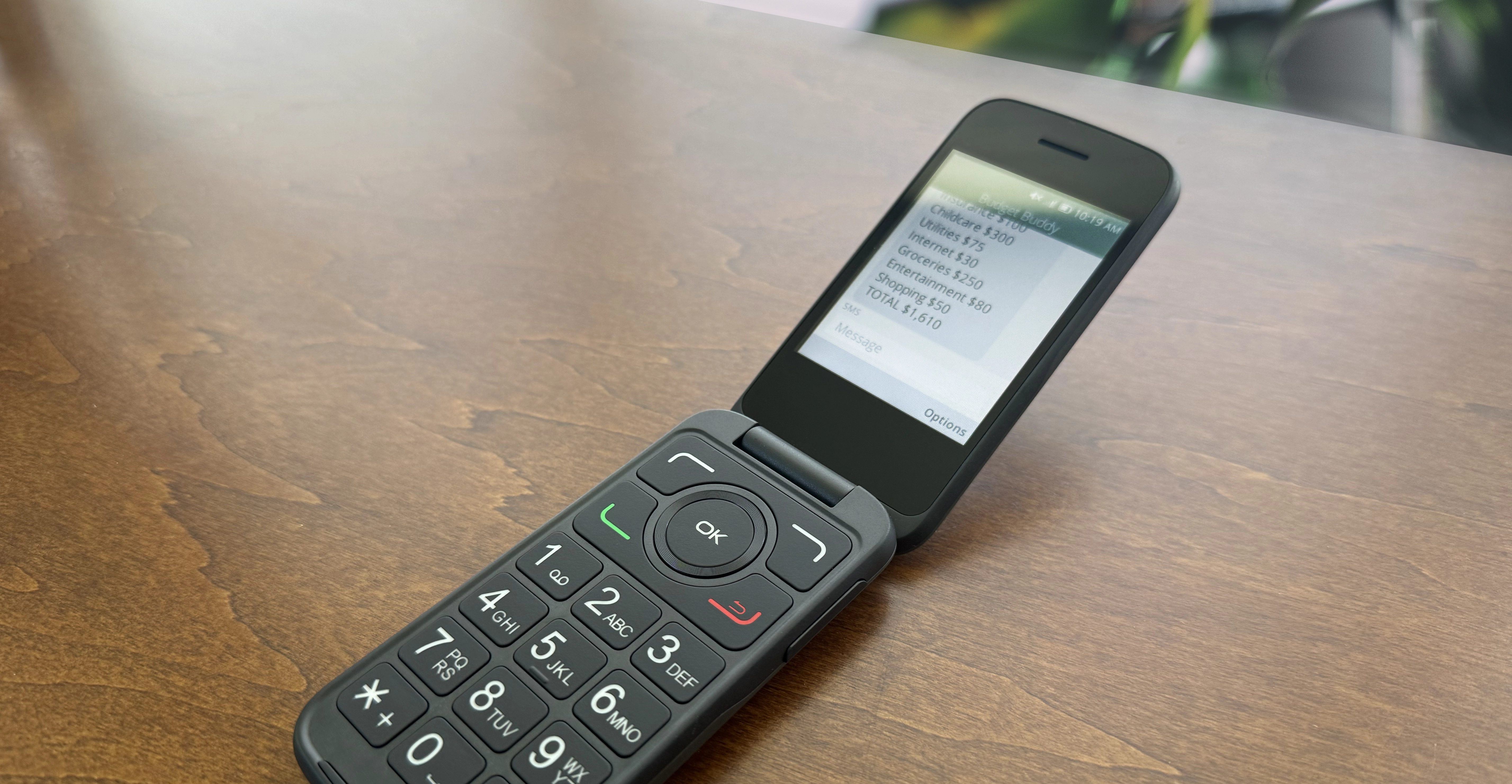 Black flip phone on wooden table with screen showing a budget breakdown message.