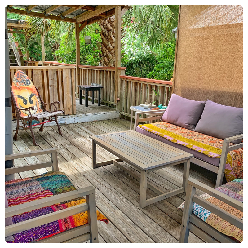 Wooden patio with a sofa, two chairs with colorful cushions, a coffee table, and green foliage outside.