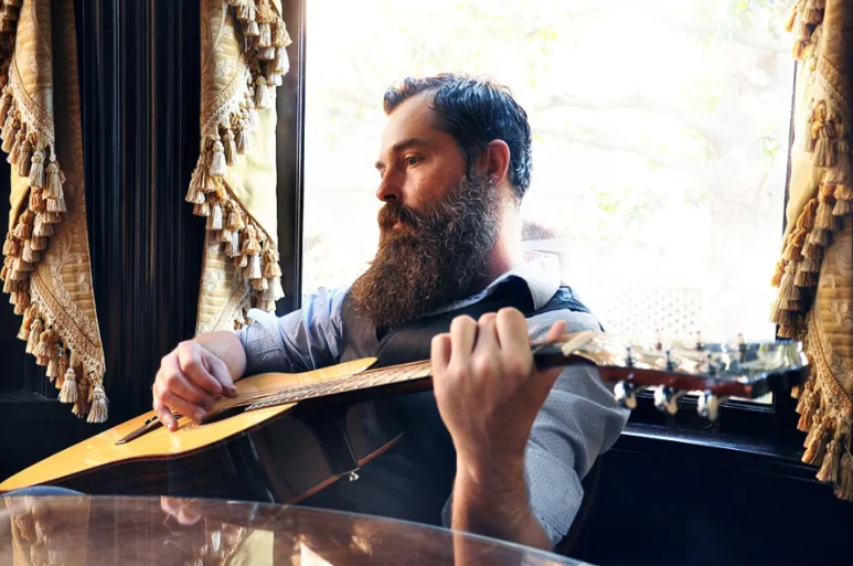 Bearded man playing acoustic guitar while sitting by a window with ornate curtains.