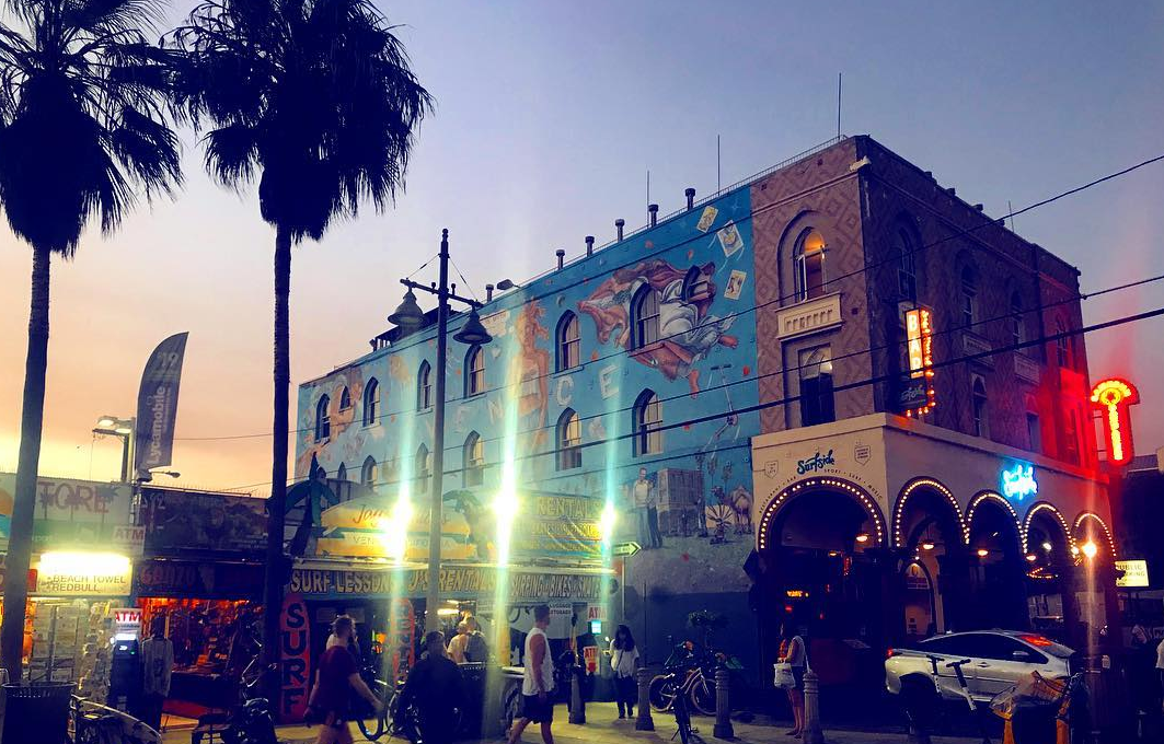 Evening street scene with palm trees, a mural on a two-story building, and people walking near shops and restaurants.