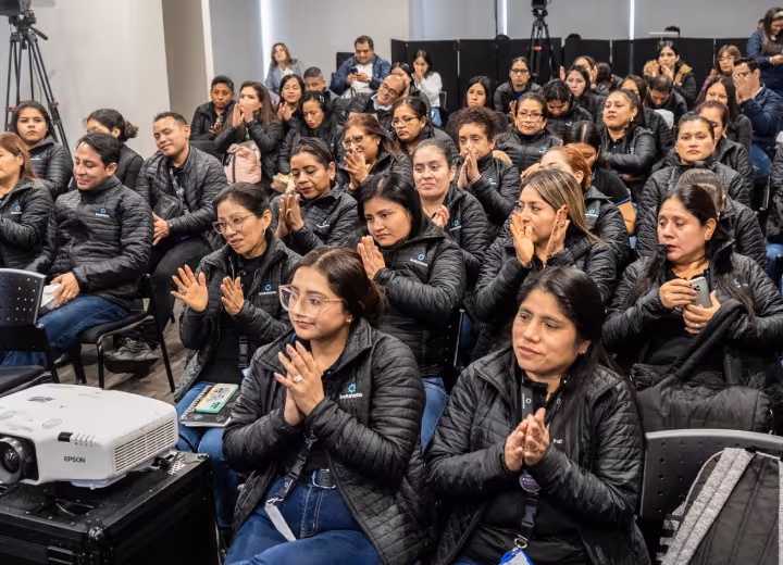 A group of people in black jackets sitting and clapping in an indoor conference or meeting room.