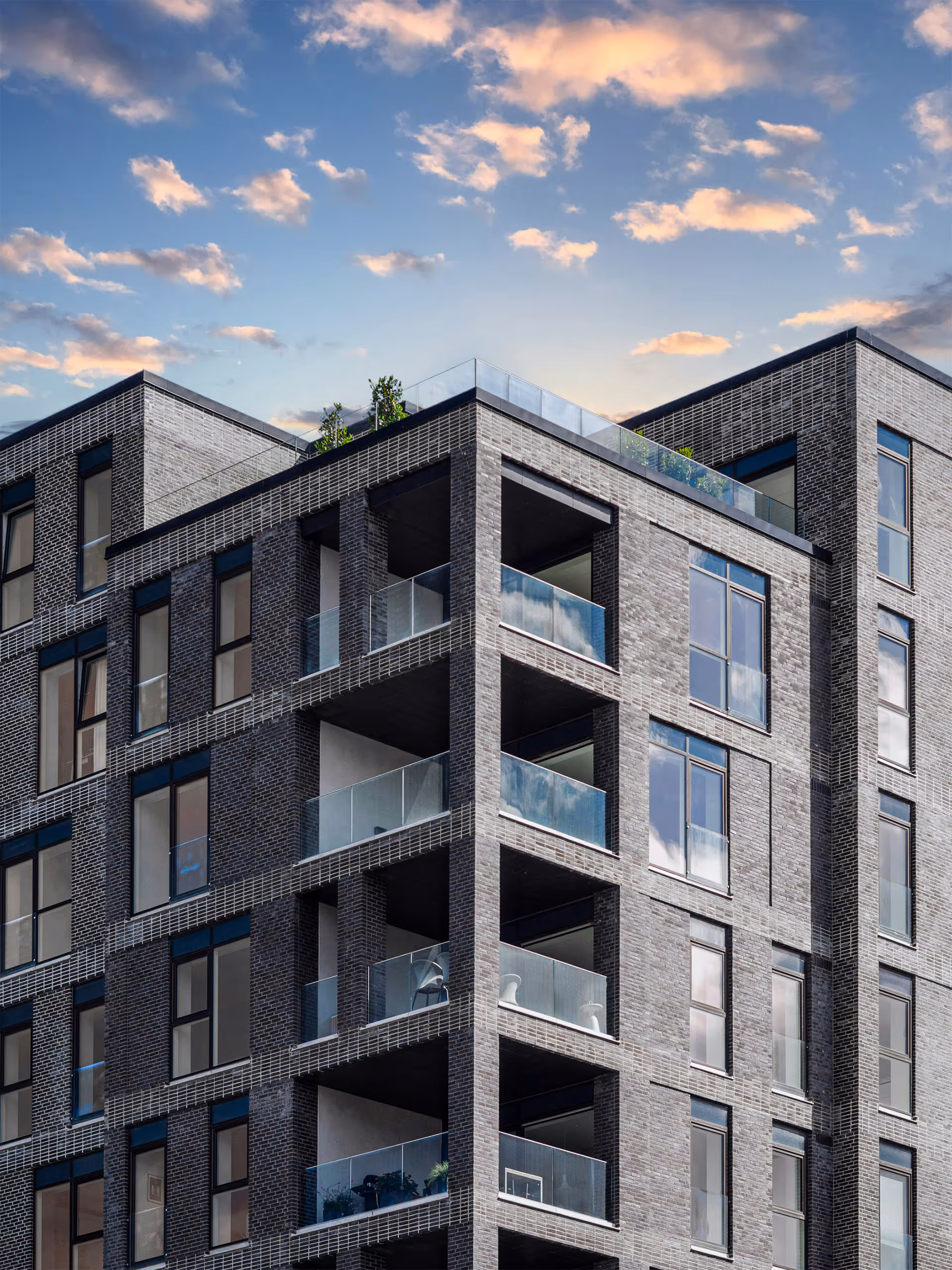 Modern multi-story building with dark brick exterior and glass balconies under a partly cloudy sky.