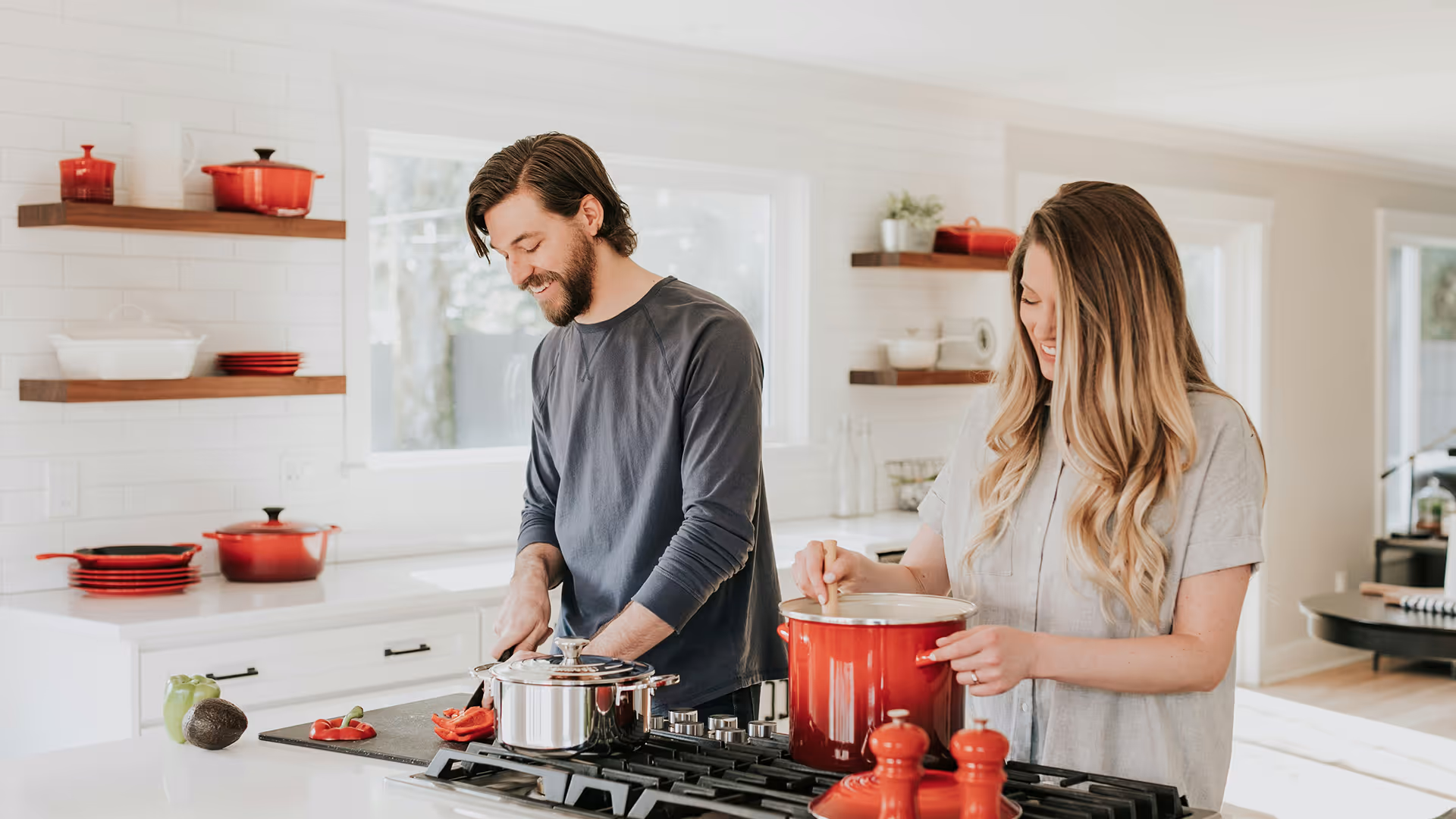Man chopping red bell pepper and woman stirring a red pot in a bright modern kitchen.