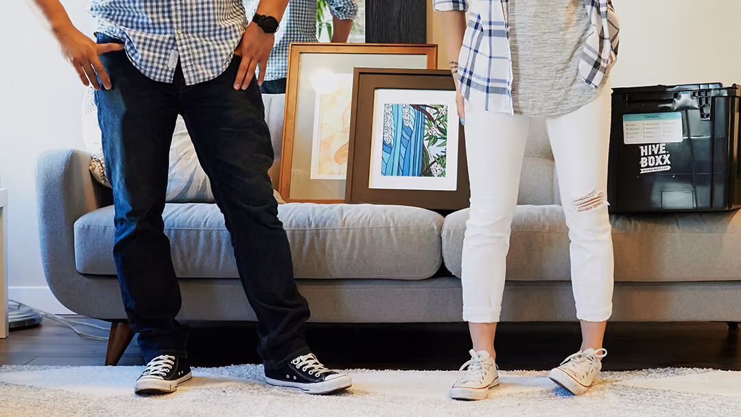 Couple standing in front of a sofa with framed art and a moving box nearby.