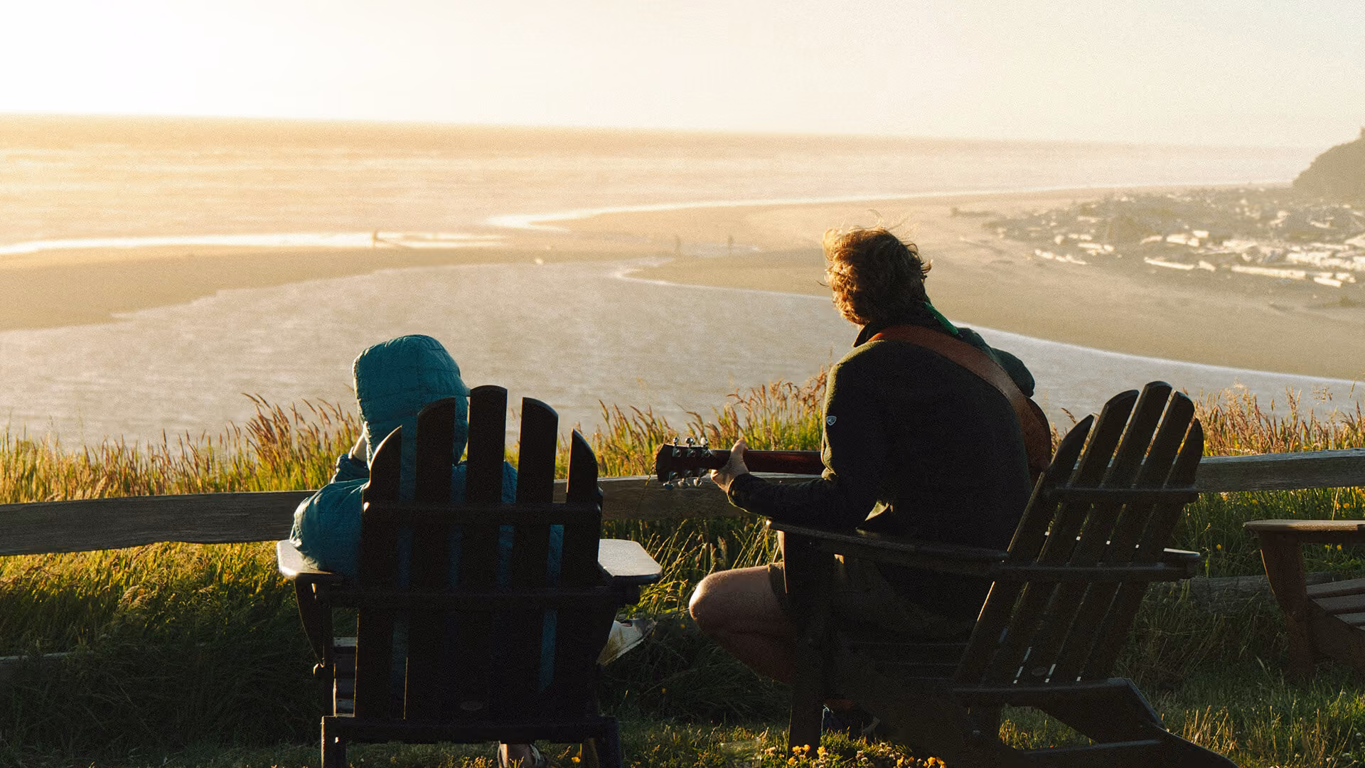 Two people sitting in wooden chairs on a hill, overlooking a beach and ocean at sunset, one plays guitar.
