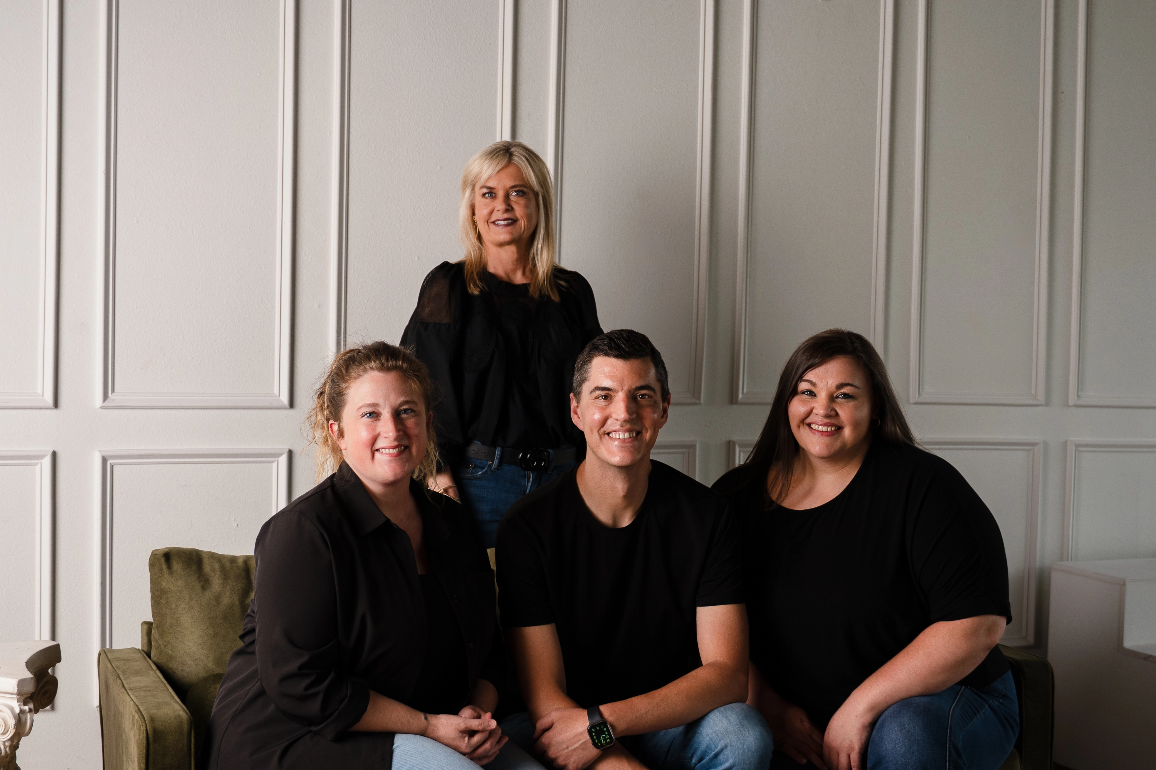 Four adults smiling, three seated on a green couch and one standing behind against paneled wall.