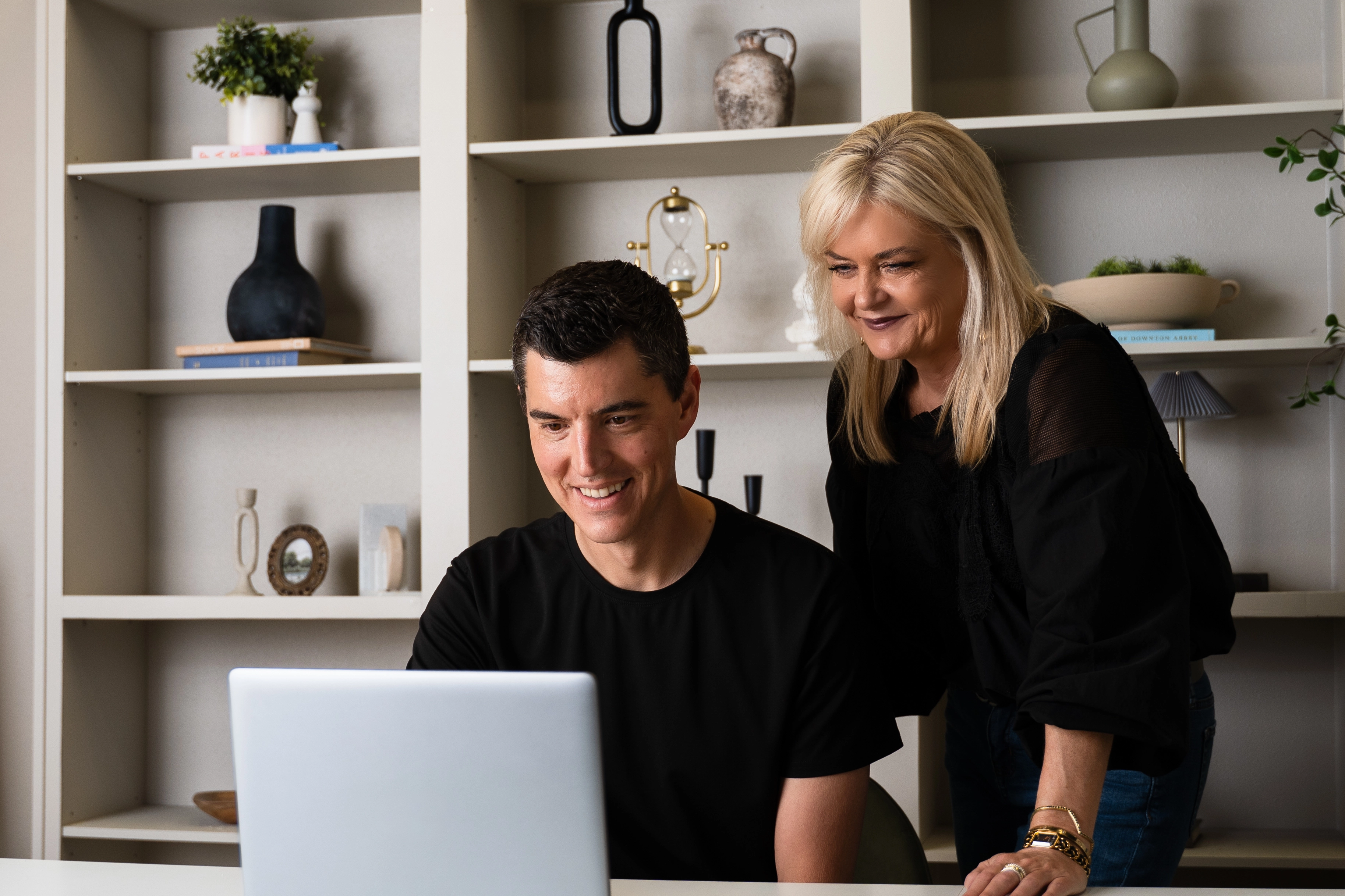 Smiling man sitting at a desk with a laptop while a woman stands beside him looking at the screen.
