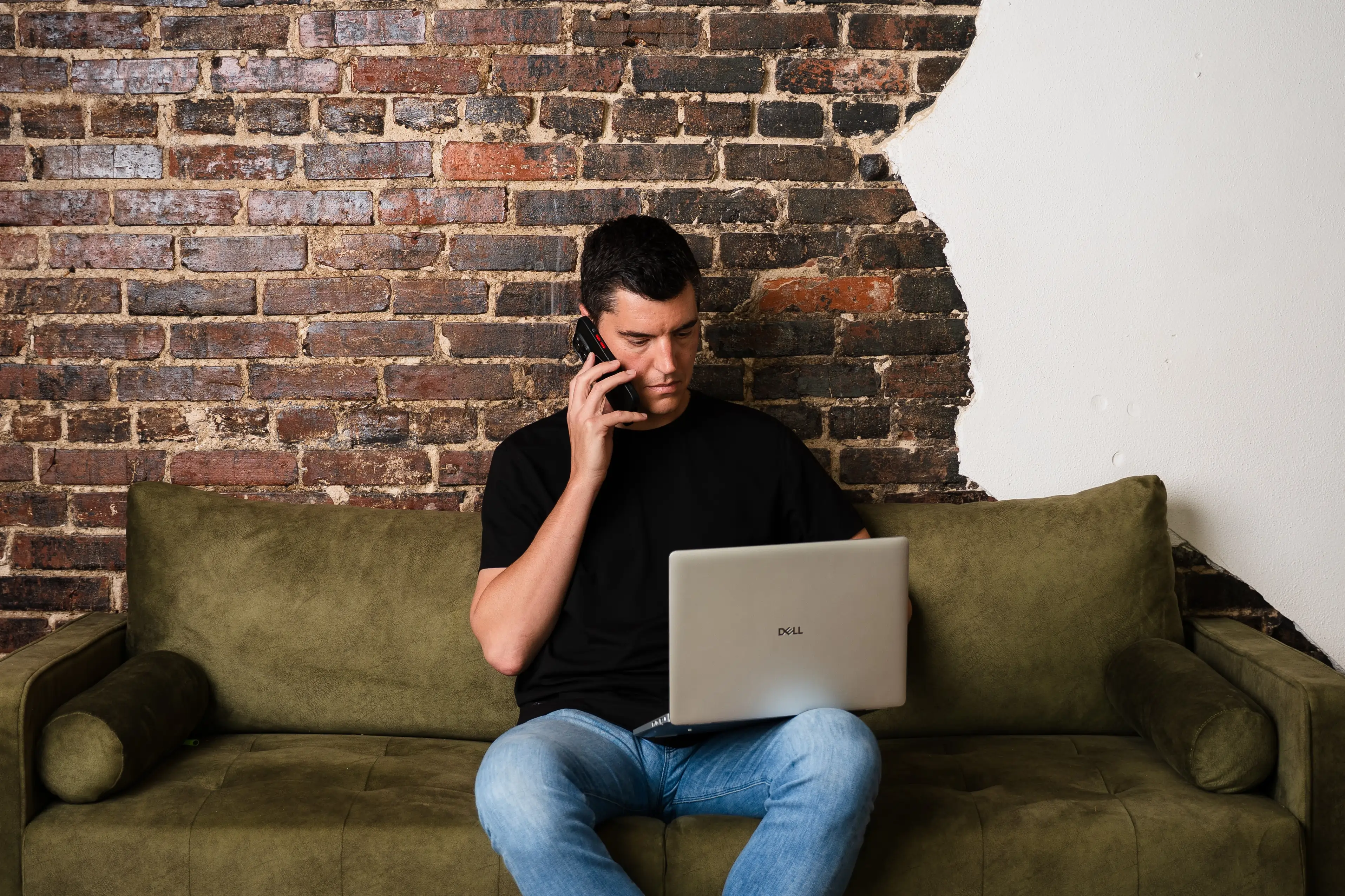 Man in black shirt using a Dell laptop and talking on phone on a green couch against a brick wall.
