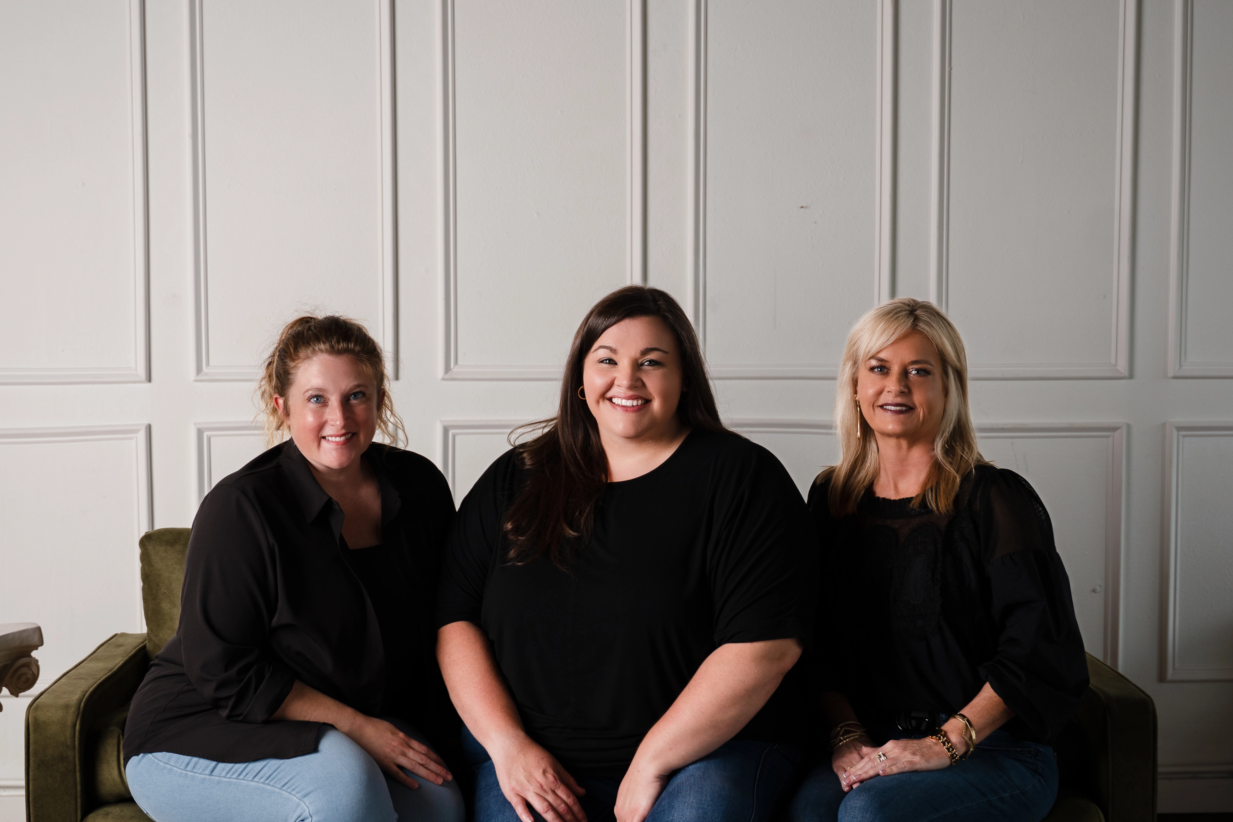 Three women in black tops sitting on a green couch against a white paneled wall.