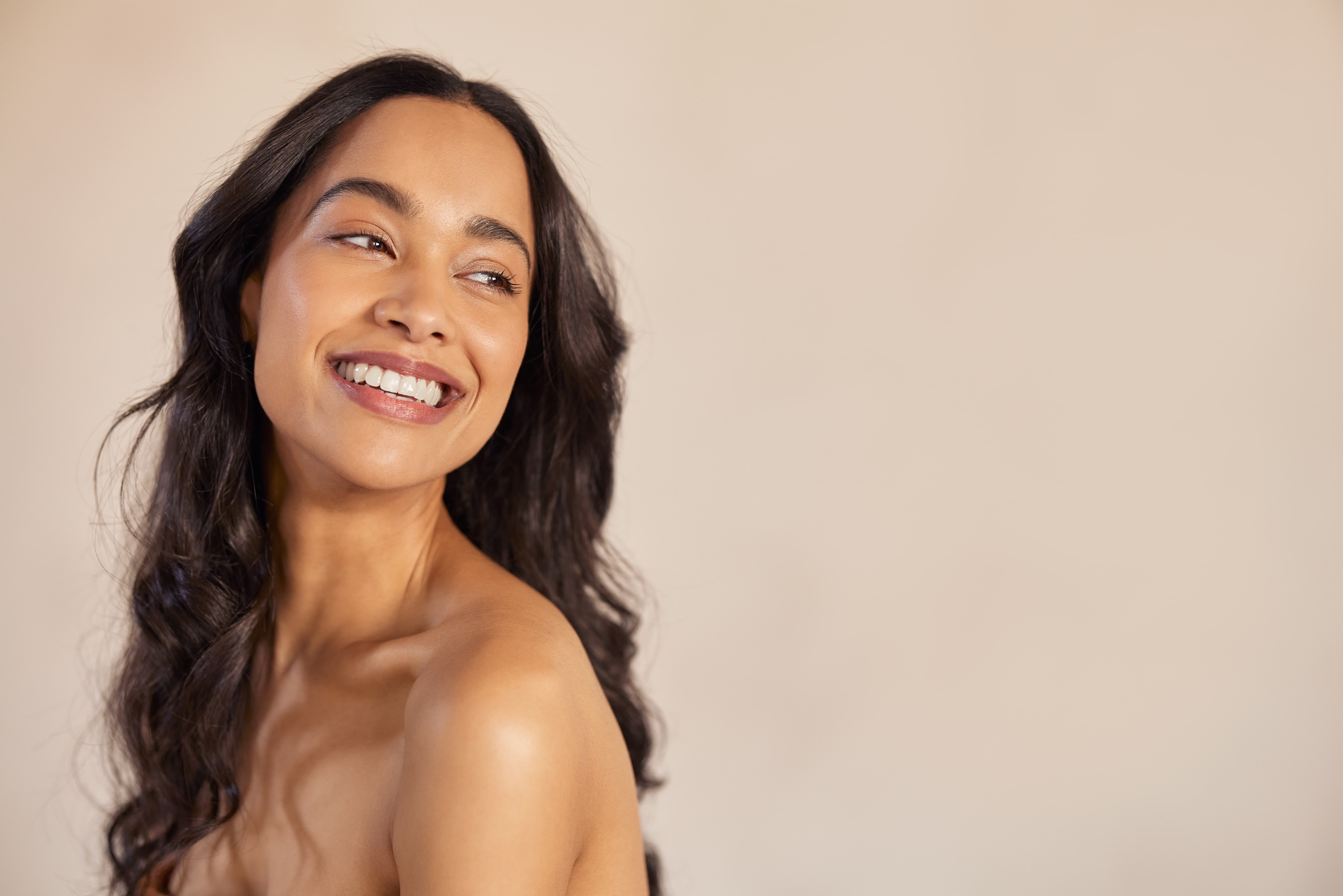 Smiling woman with long wavy dark hair looking to her left on a beige background.