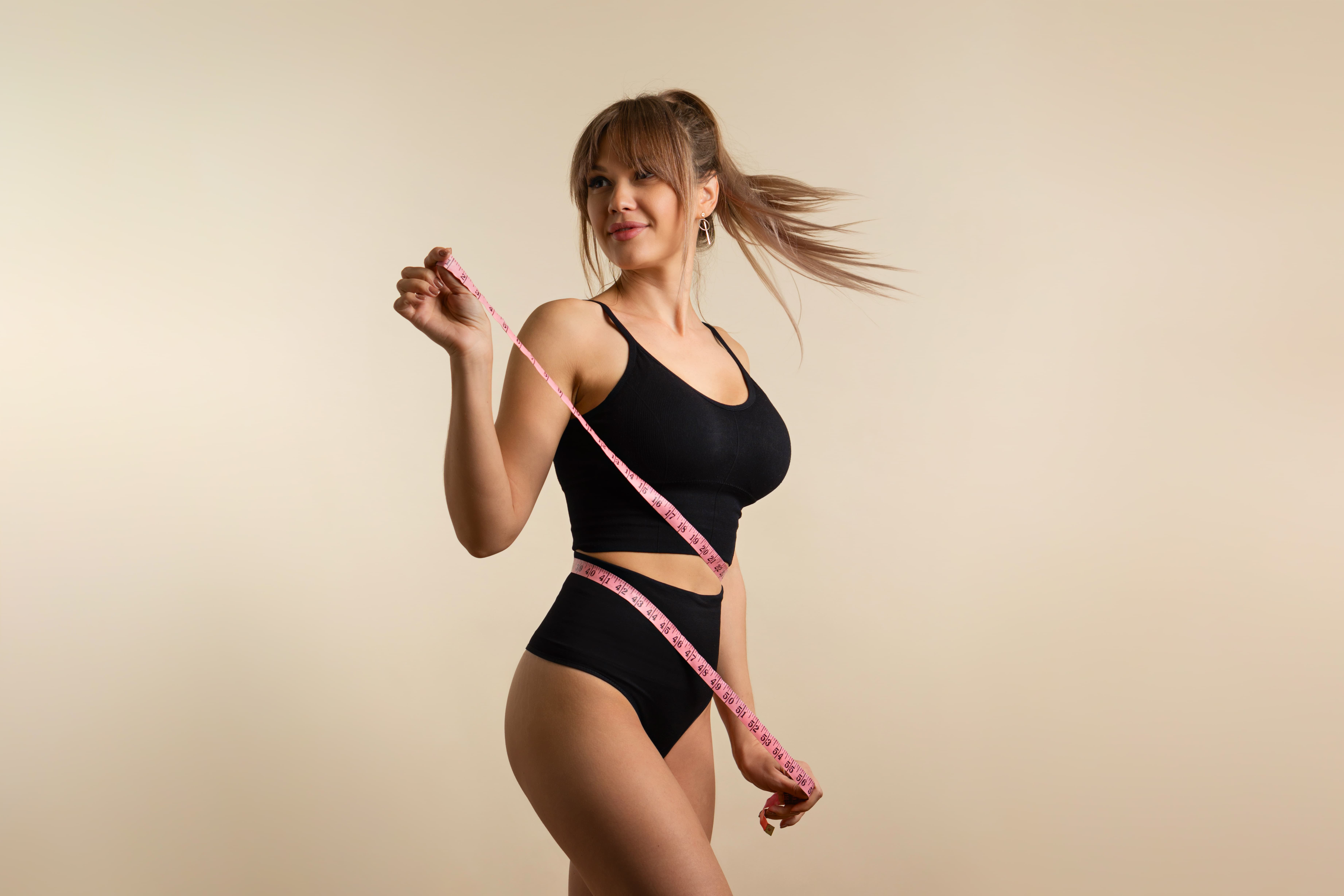 Woman in black sportswear measuring waist with pink tape measure against beige background.