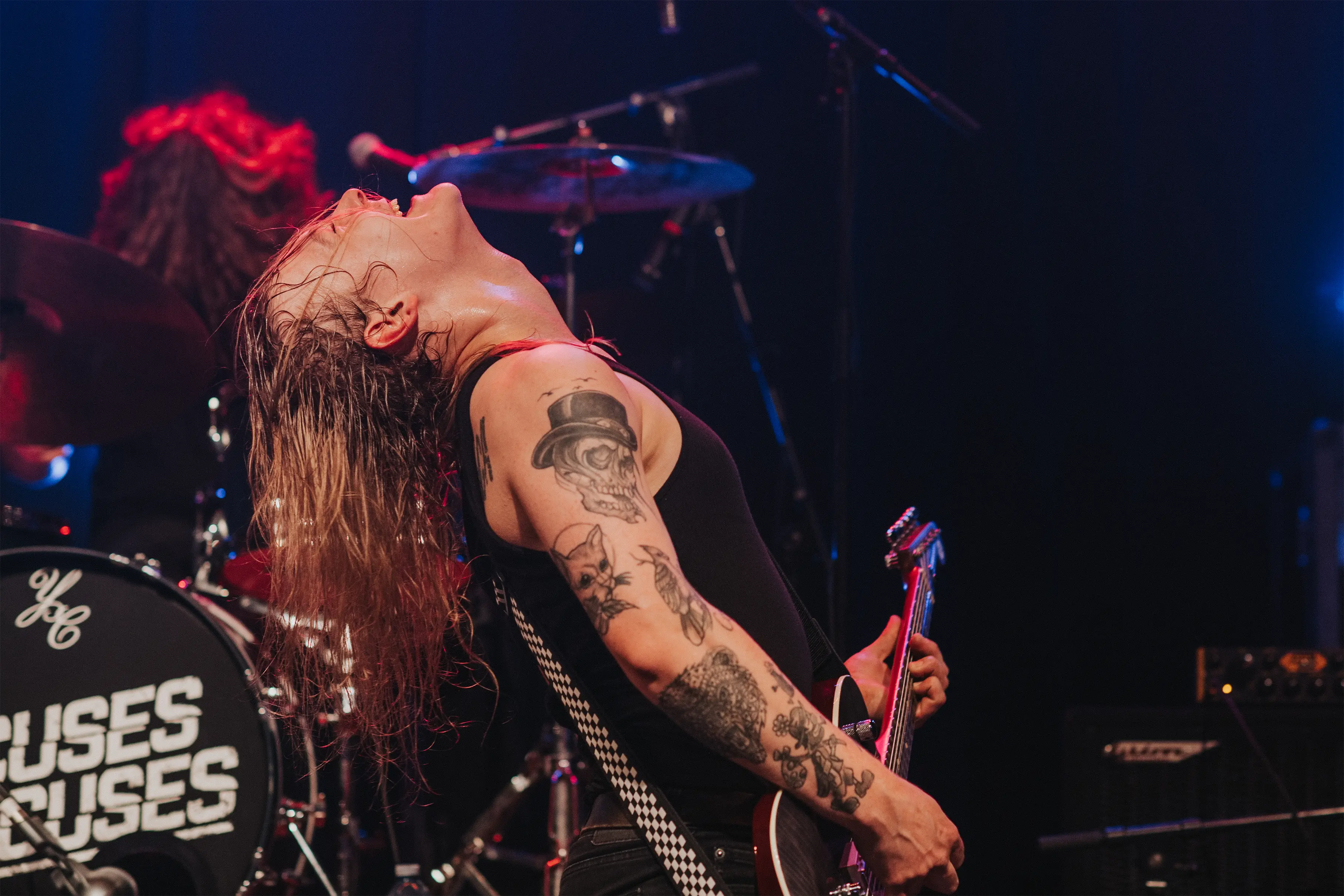 Long-haired guitarist with multiple tattoos on arm, playing electric guitar energetically on stage with head tilted back and eyes closed, a drummer visible in the background near a drum kit labeled 'Excuses'.