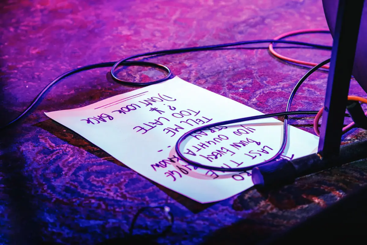Setlist paper on a patterned stage floor with some black cables and a microphone visible around it, illuminated by purple and blue stage lights.