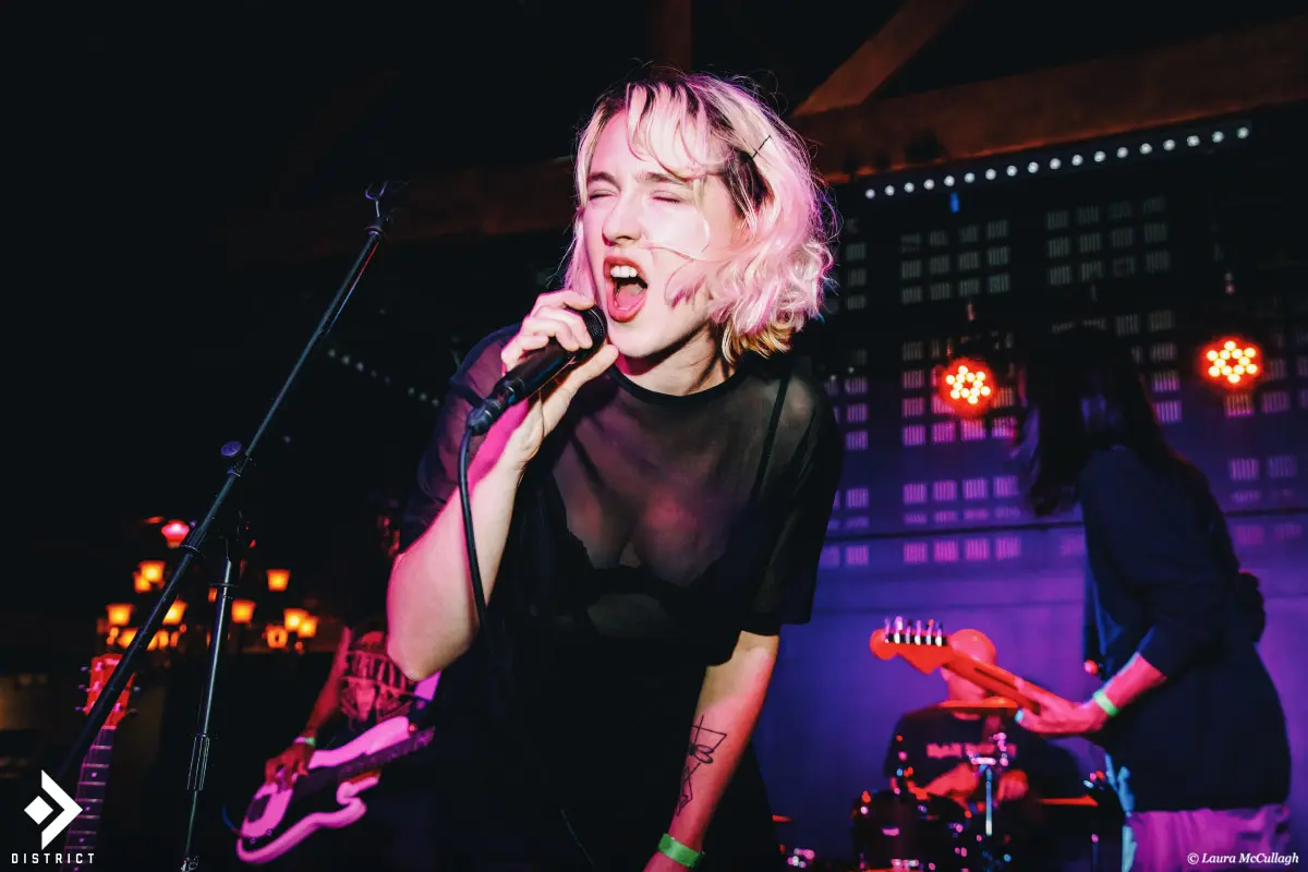 Female singer with short wavy blonde hair singing passionately into a microphone on stage, wearing a sheer black top, with band members playing guitar and drums in purple and red stage lighting.
