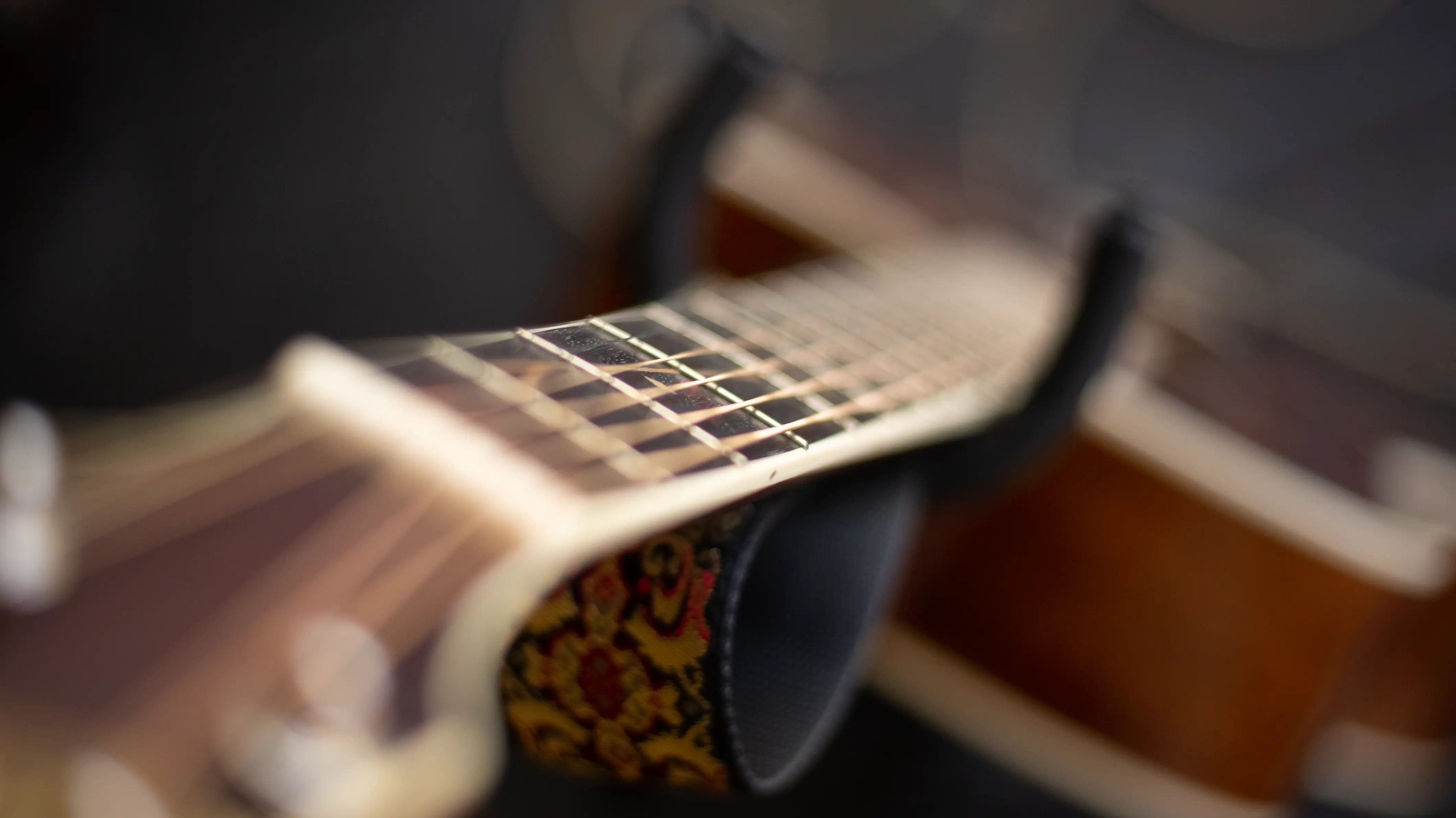 Close-up view of an acoustic guitar neck and fretboard with metal strings and a decorative strap underneath.