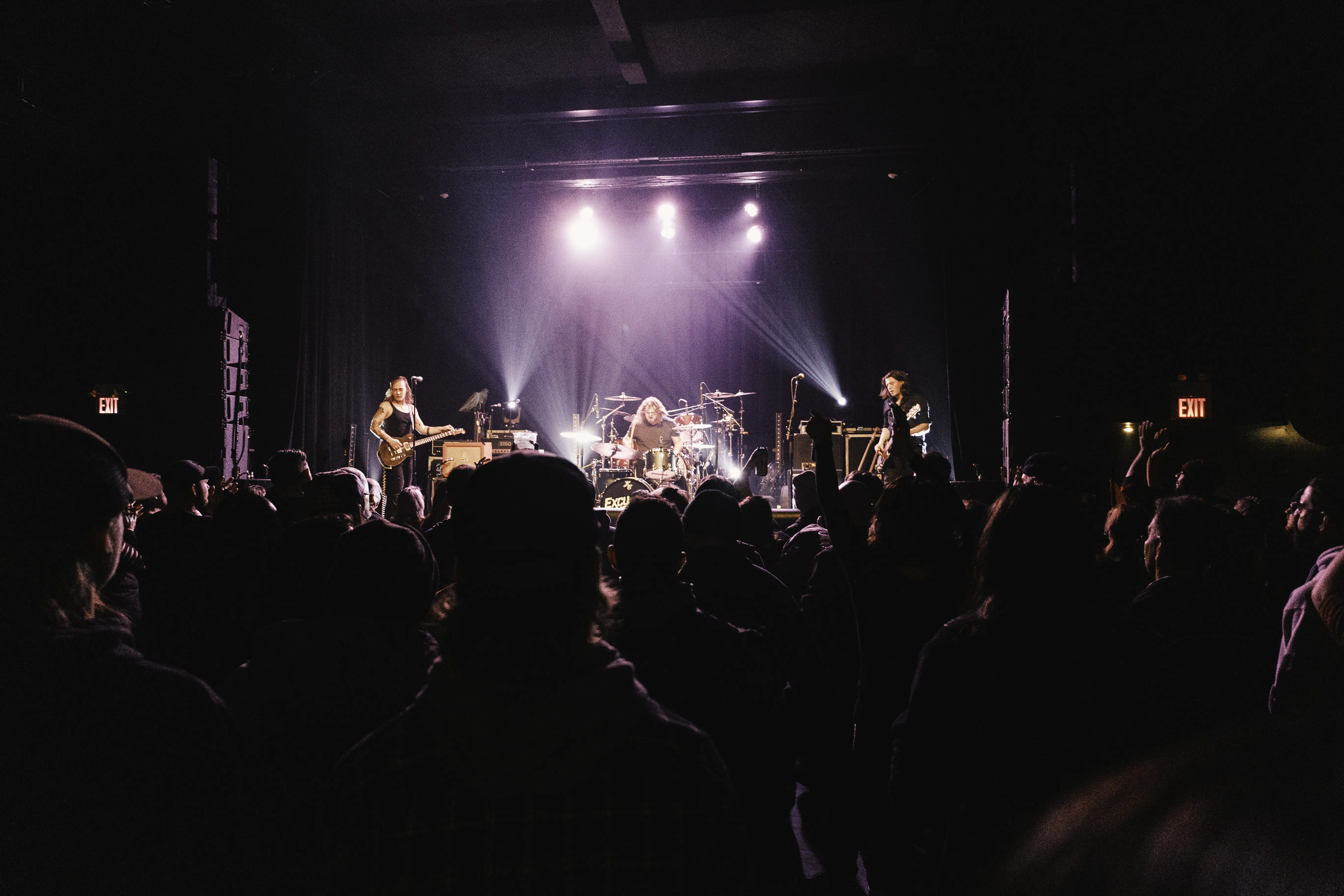 Live concert scene with a three-piece band performing on stage under purple and white spotlights and a crowd watching in a dark venue.
