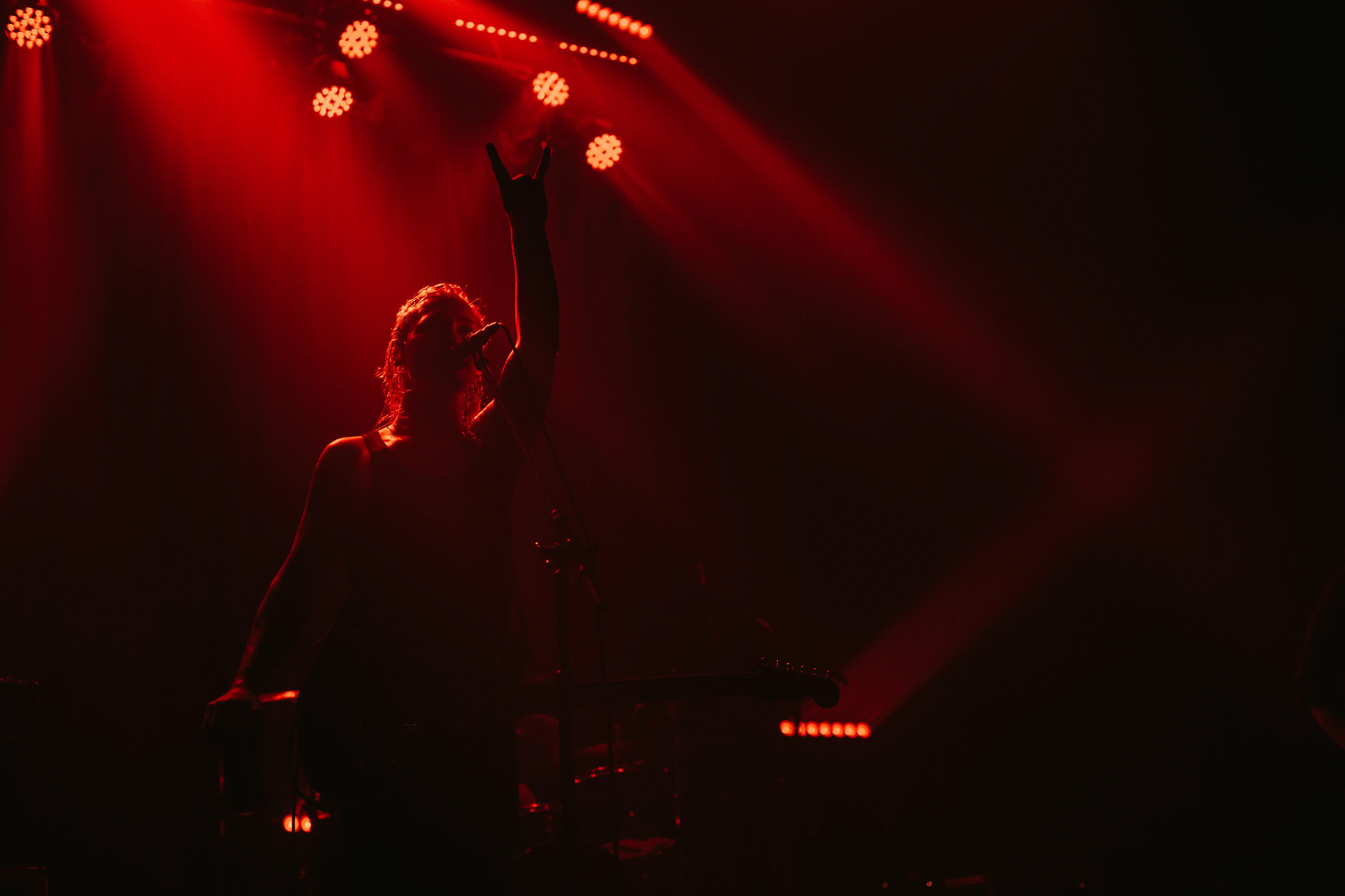 Silhouetted musician on stage with guitar, raising one hand making the rock and roll gesture under red stage lights.