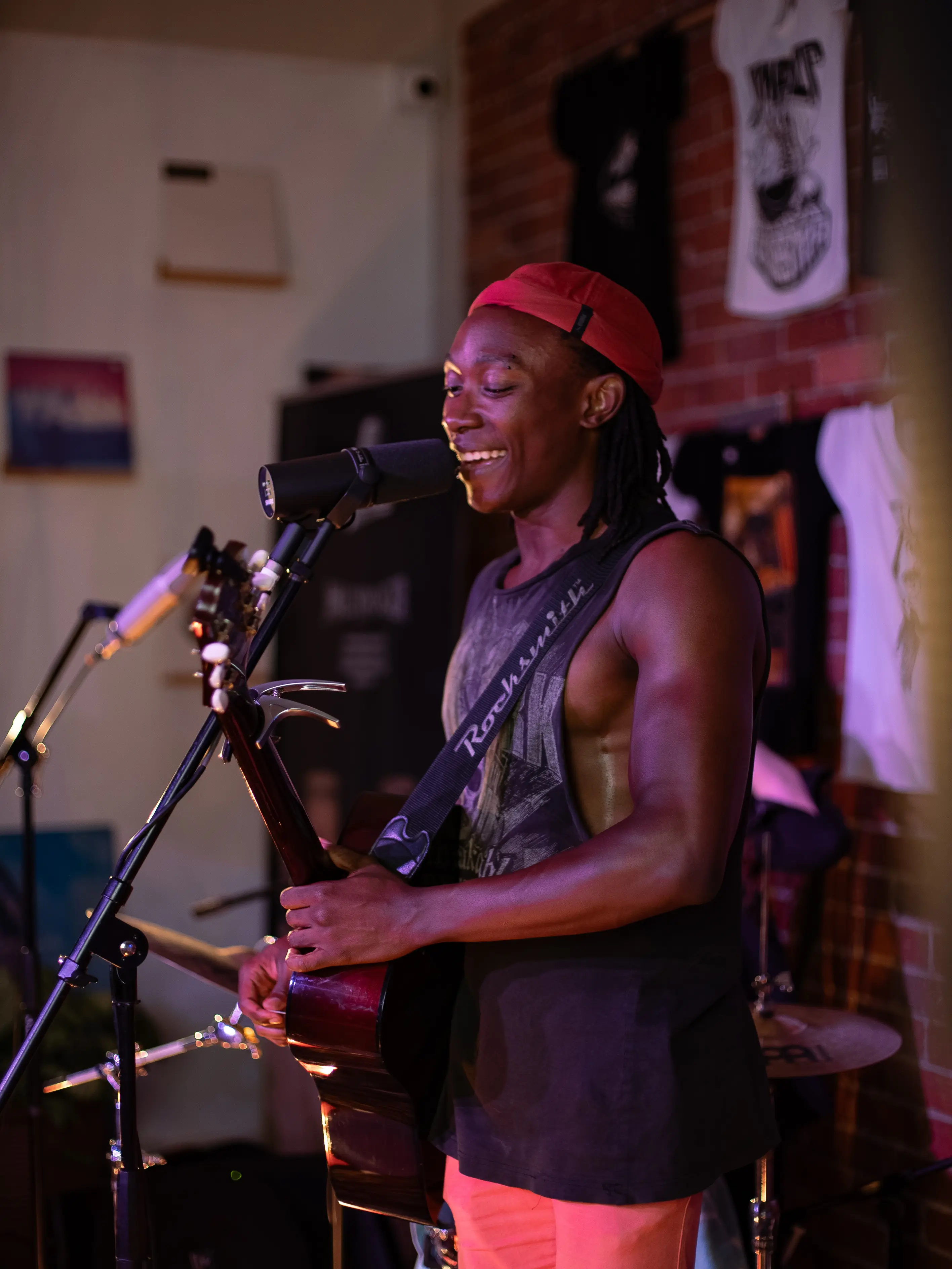 Musician wearing a red cap backward, sleeveless black shirt, and pink pants playing an acoustic guitar and singing into a microphone on a stand indoors with dim lighting and band equipment in the background.