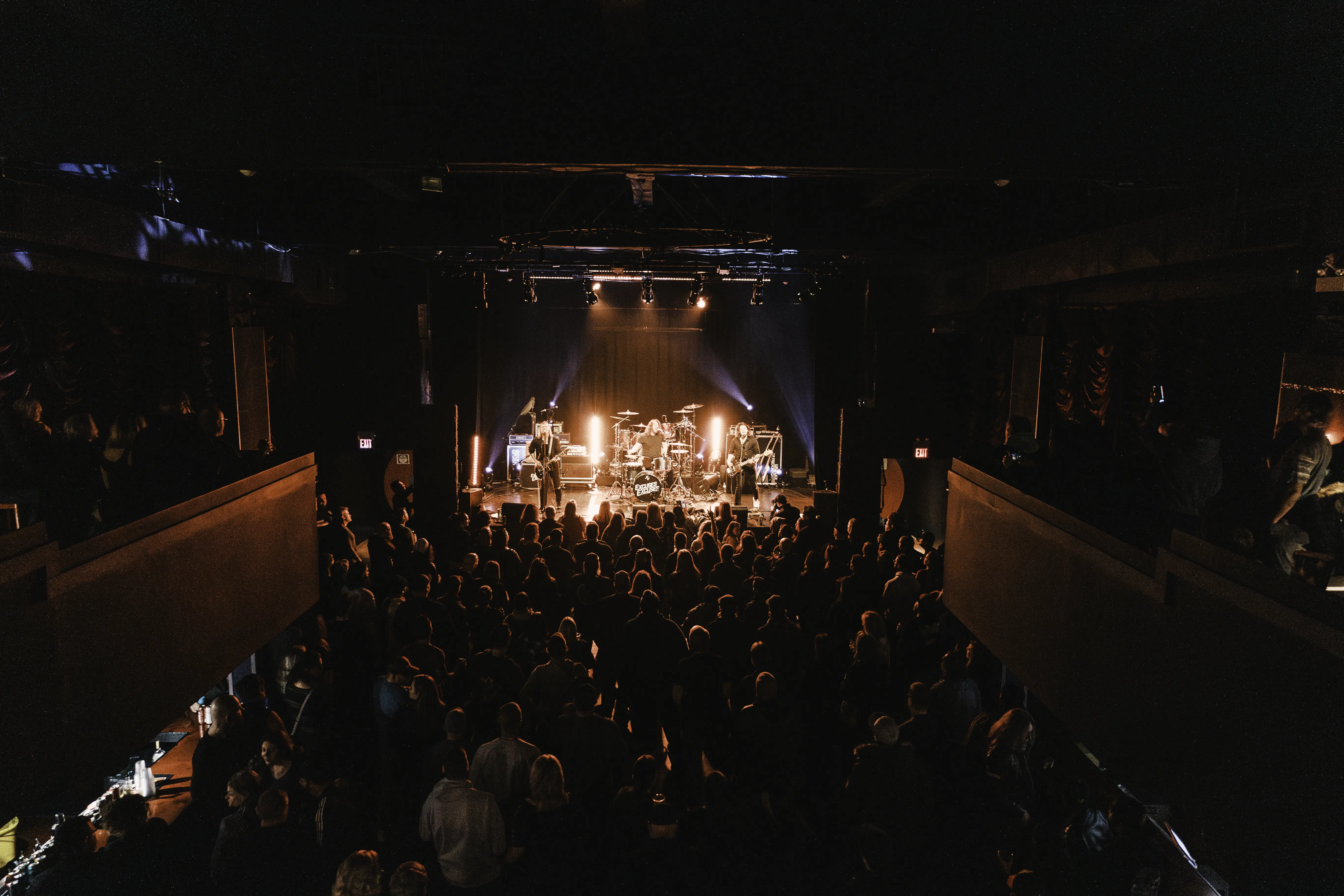Crowded indoor concert venue with a three-member band performing on stage under orange and blue lights, audience watching.