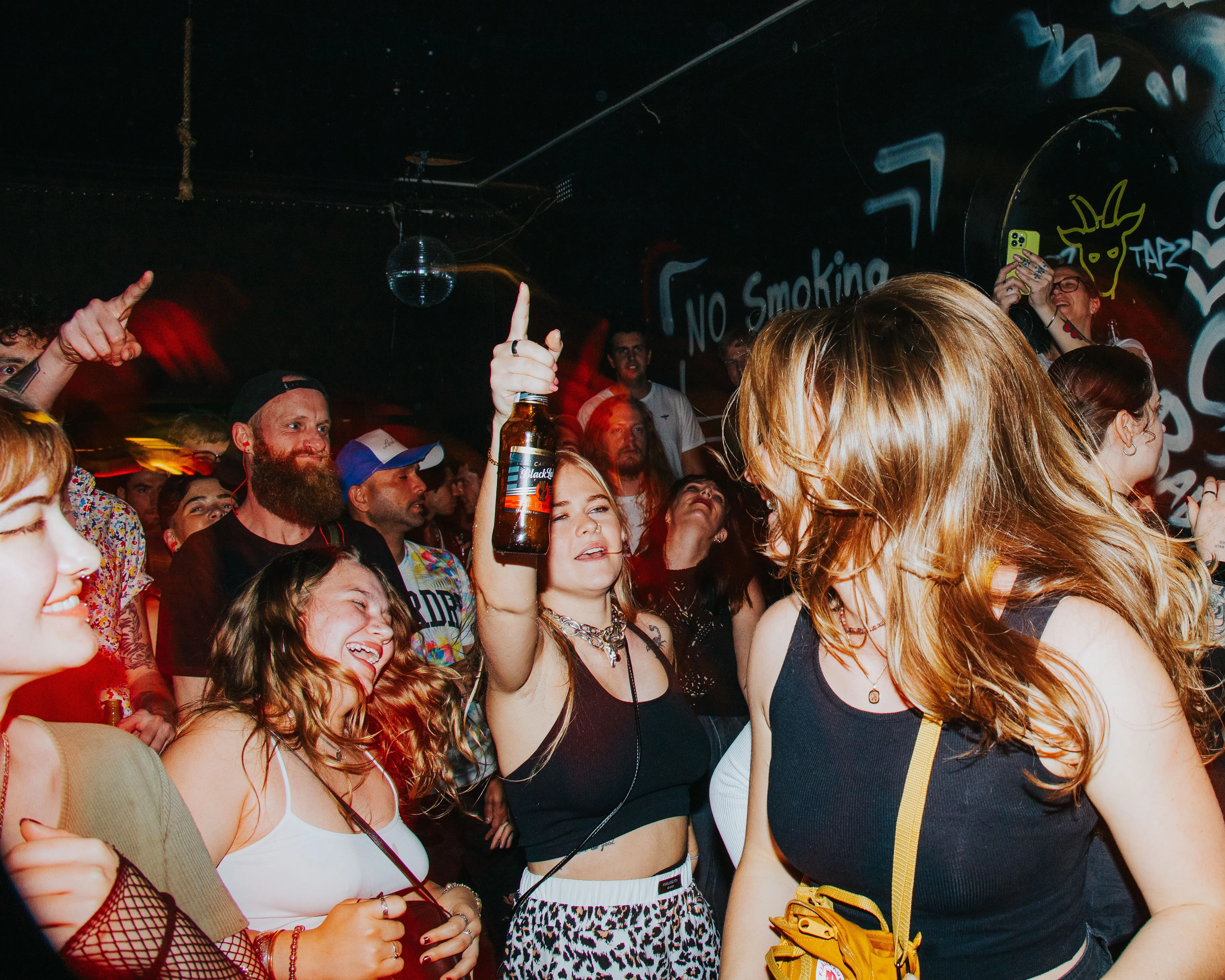 A lively group of young people dancing and enjoying a crowded party or nightclub with dim lighting and graffiti walls, one woman holding a bottle up in celebration.