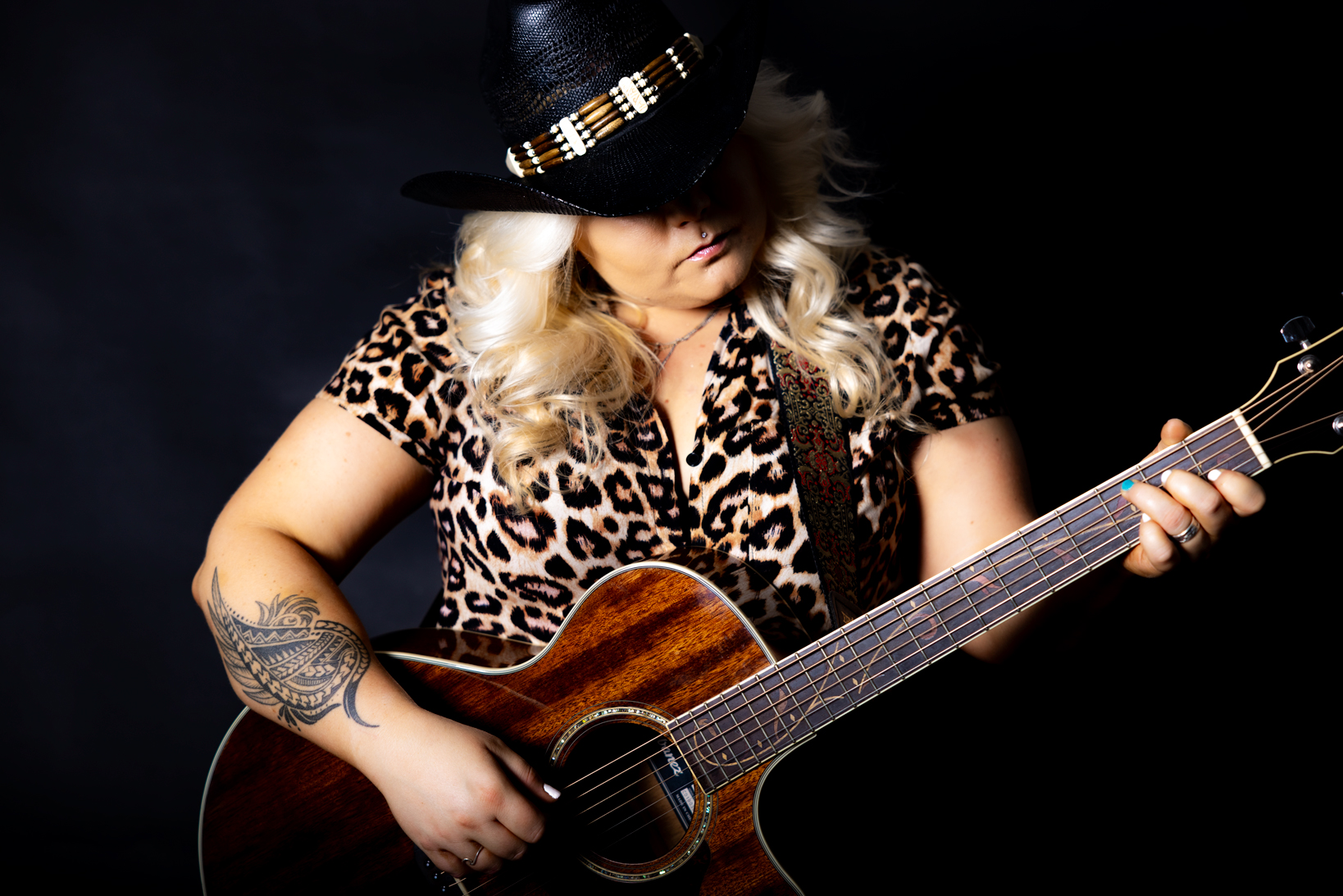 Woman with long blonde hair wearing a black hat with beads and a leopard print shirt playing an acoustic guitar, showing a detailed tattoo on her left arm against a dark background.