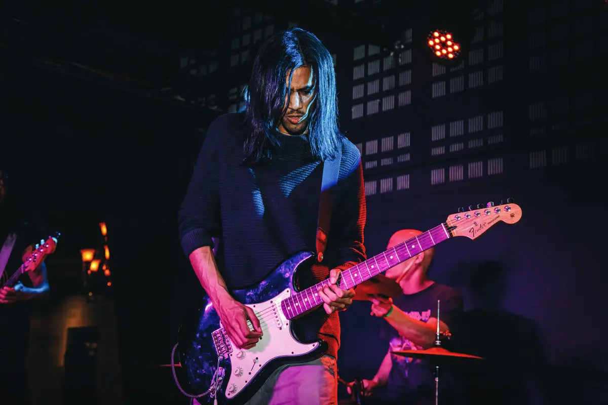 Musician with long hair playing an electric guitar on stage during a live performance, with a drummer visible in the background.