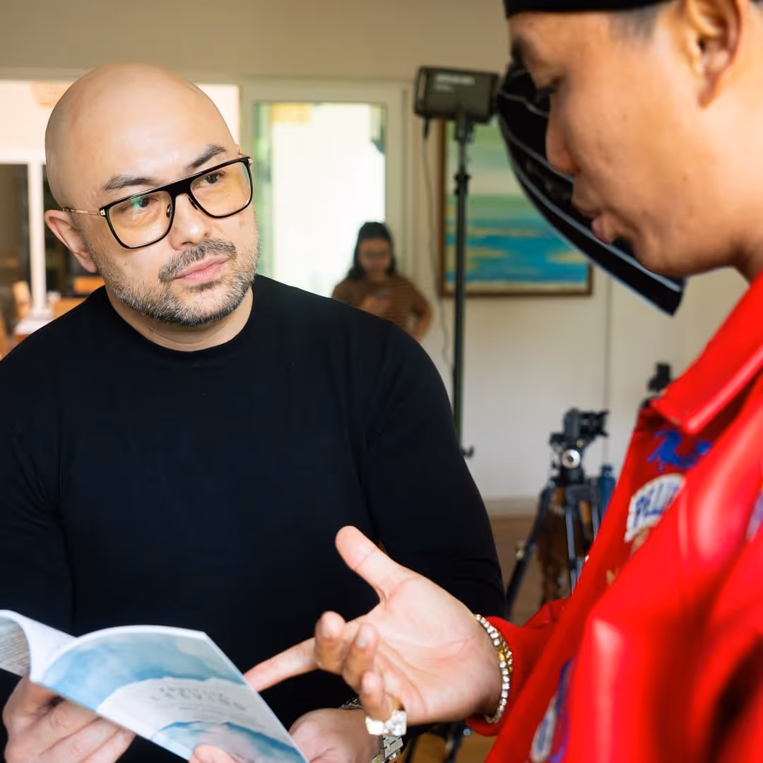 Two men engaged in a discussion while looking at a booklet, one wearing glasses and a black shirt, the other in a red jacket.