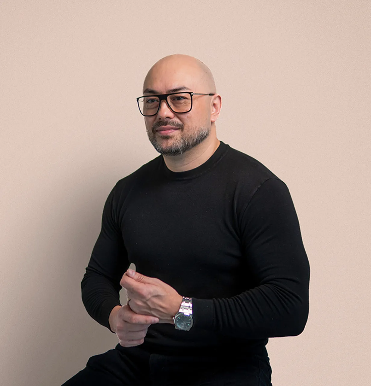 Paul Quinn wearing a black long-sleeve shirt and a wristwatch sitting against a beige background.