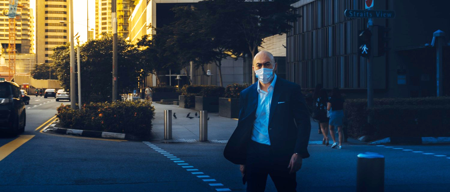 Paul Quinn in sunglasses and dark suit walking on a city sidewalk near a street with cars and greenery.