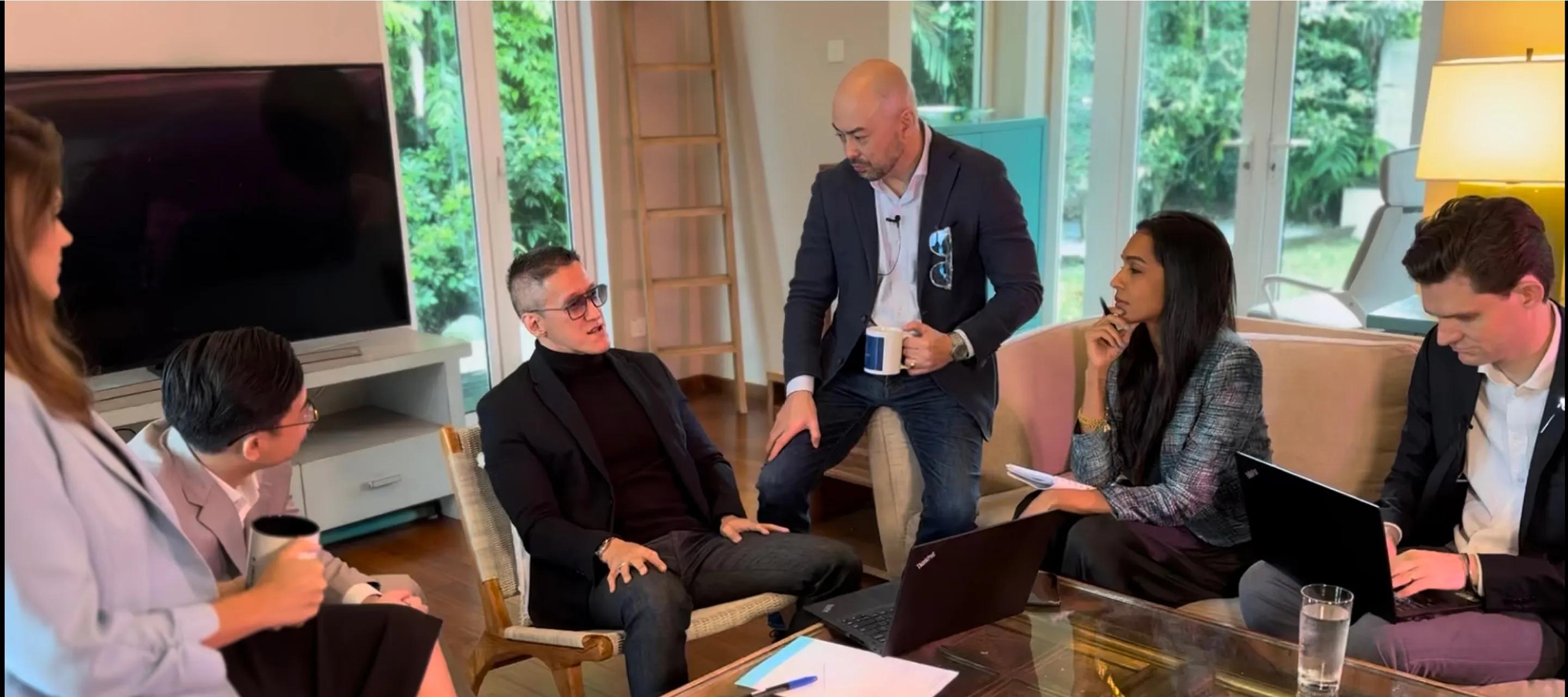 Two people sitting in green chairs in a bright room; a woman wearing a blue and white striped shirt and white skirt, and a man with glasses in a navy blazer and jeans.
