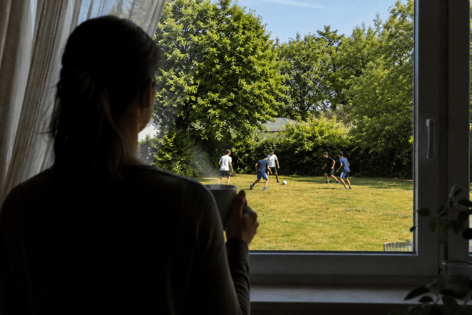 Mother holding a steaming cup of coffee and watching teenagers play football through the window.