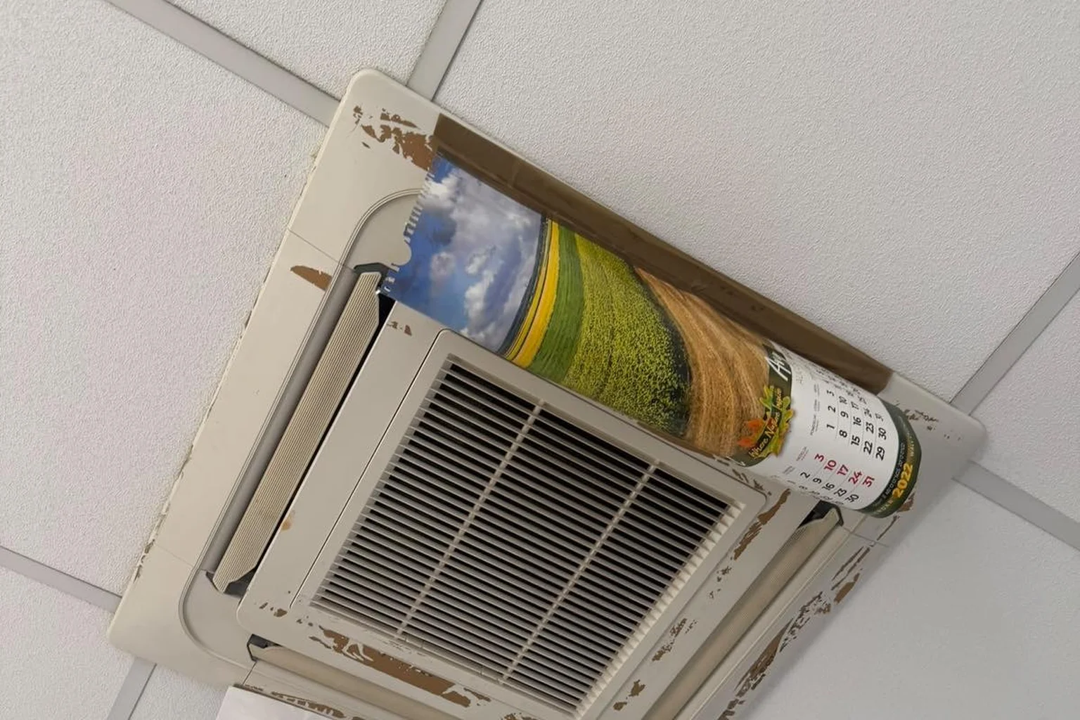 A ceiling air-conditioning unit with peeling edges, held in place with brown tape and a rolled-up calendar.