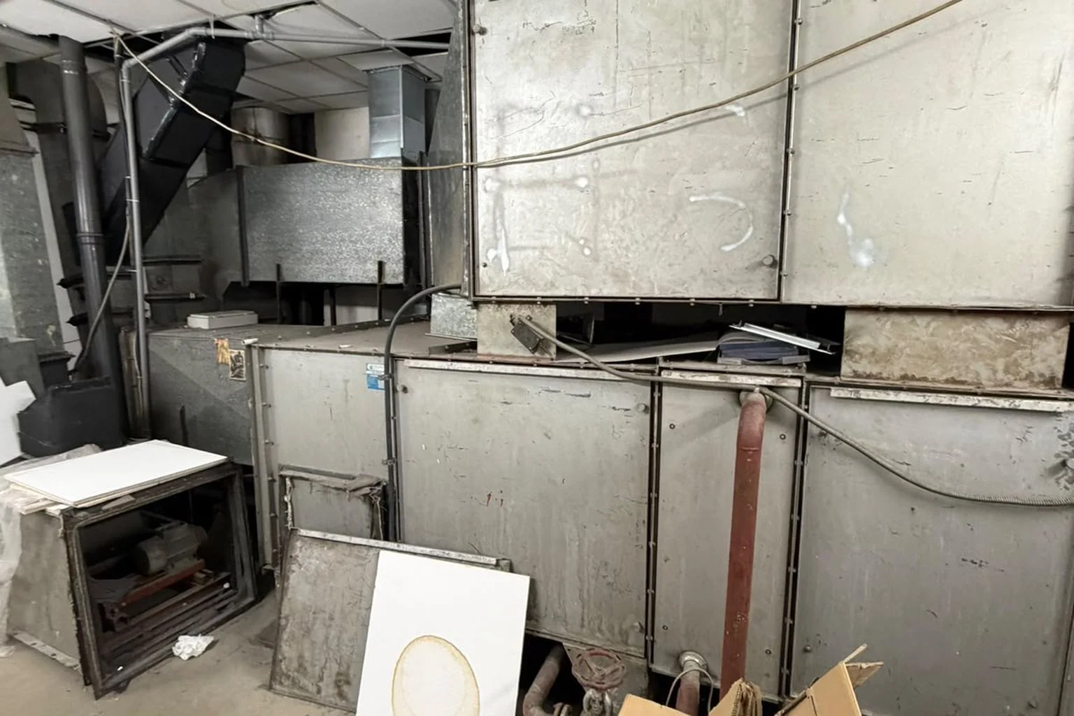 Large, dented metal air-conditioning units in a basement plant room, with pipes, loose panels and cardboard on the floor.