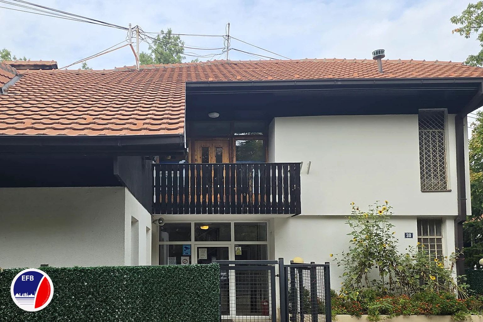 A white two-storey villa with a red tiled roof and a dark wooden balcony behind a green hedge fence, with the French School of Belgrade logo in the corner.