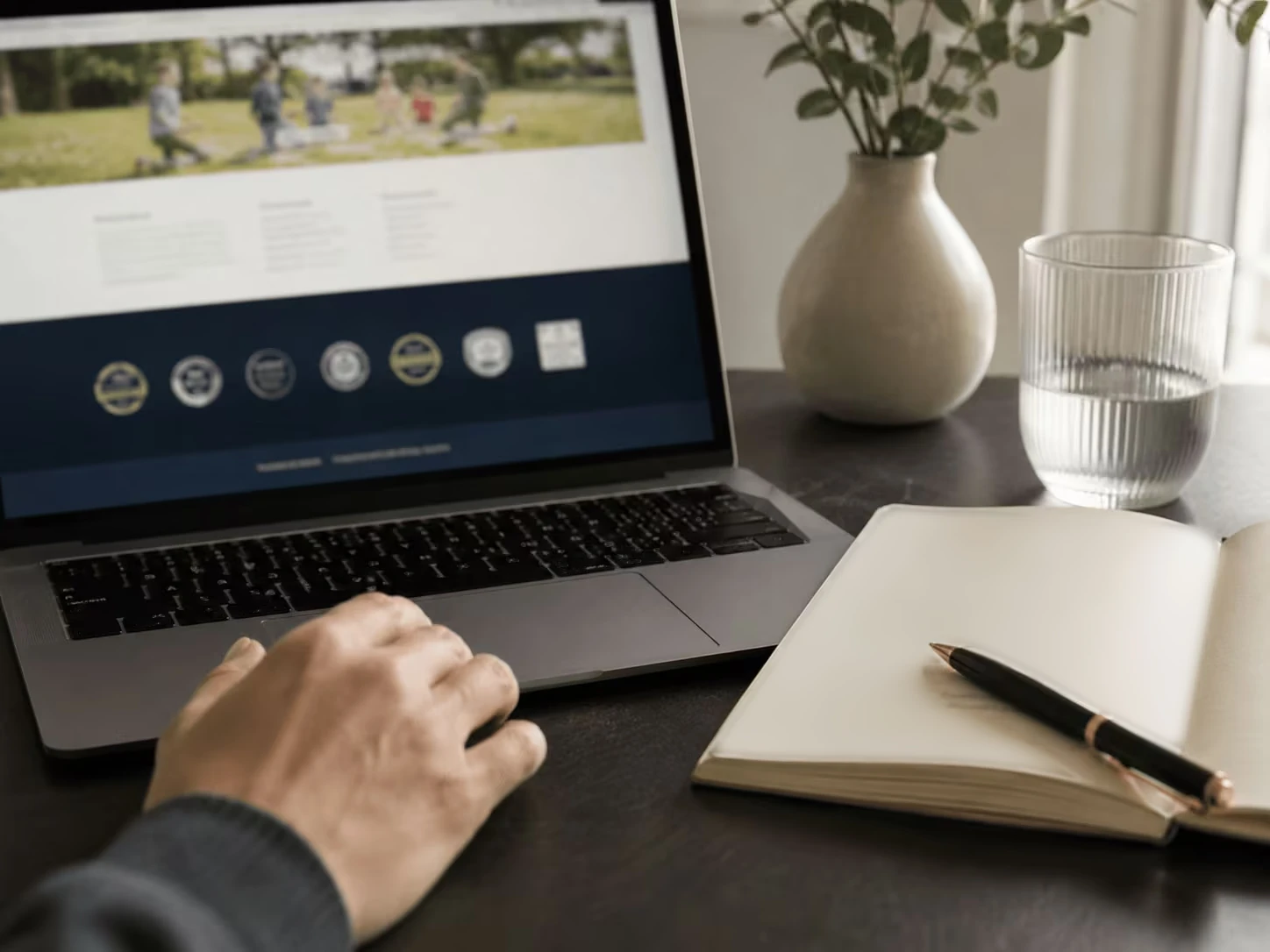 A hand rests on a laptop showing a school website with a row of accreditation emblems, beside an open notebook.