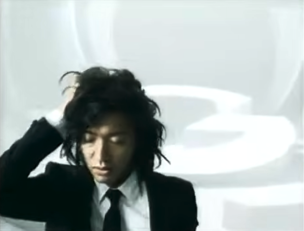 Takuya Kimura in a black suit and skinny tie, one hand pushed into his hair, framed off-centre against a white background
