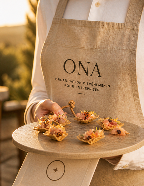 Person wearing beige apron with ONA logo holding a round tray with six elegant appetizers topped with ham and garnishes.