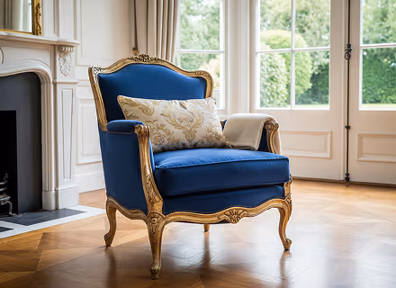 Elegant blue and gold armchair with a patterned pillow and folded blanket in a bright room.