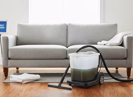 Lichte grijze bank met een stoomreiniger gevuld met vuil water ervoor en witte handdoeken ernaast.