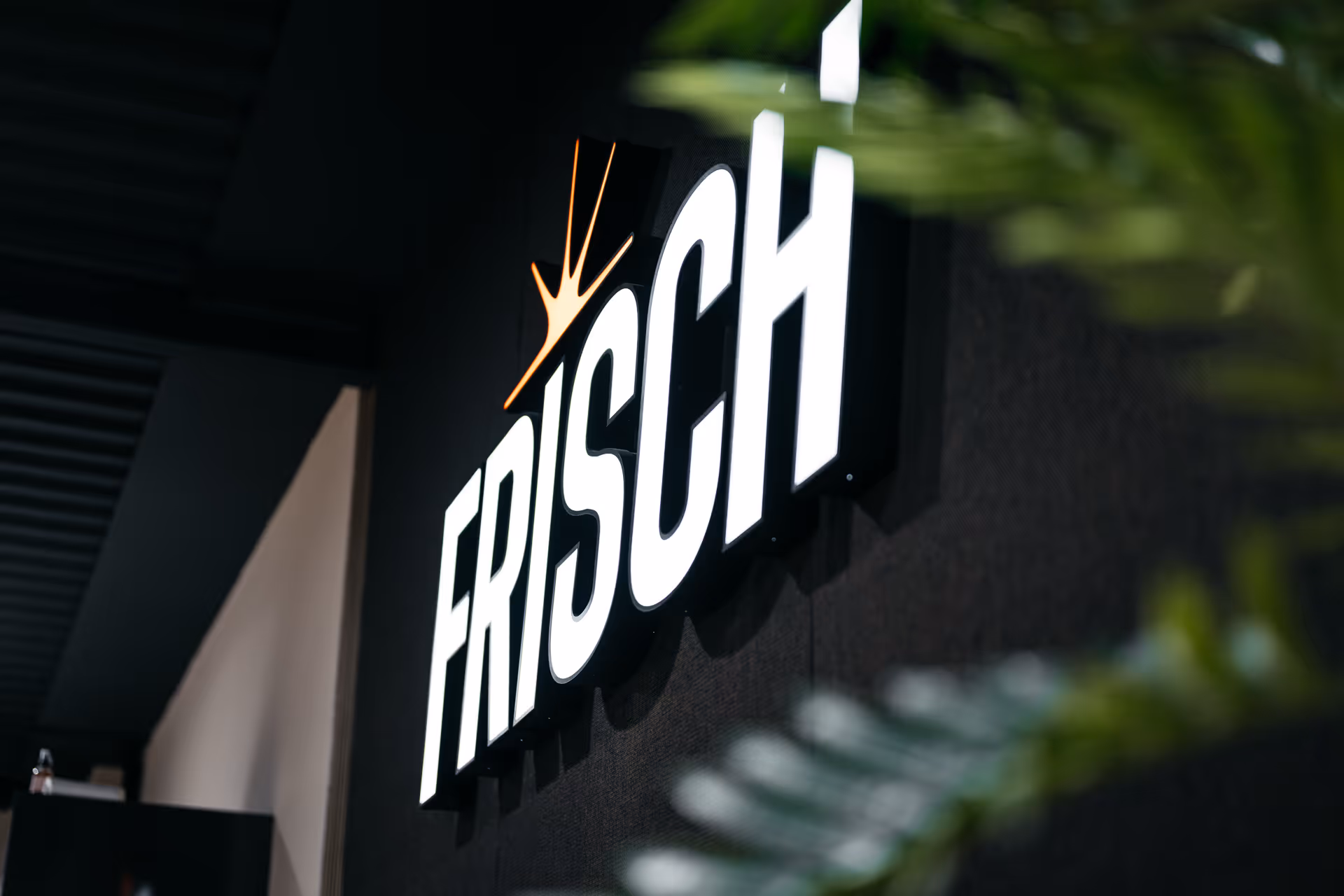 Verlichte FRISCH-sign met oranje kroon op een zwarte muur, deels bedekt door groene bladeren.
