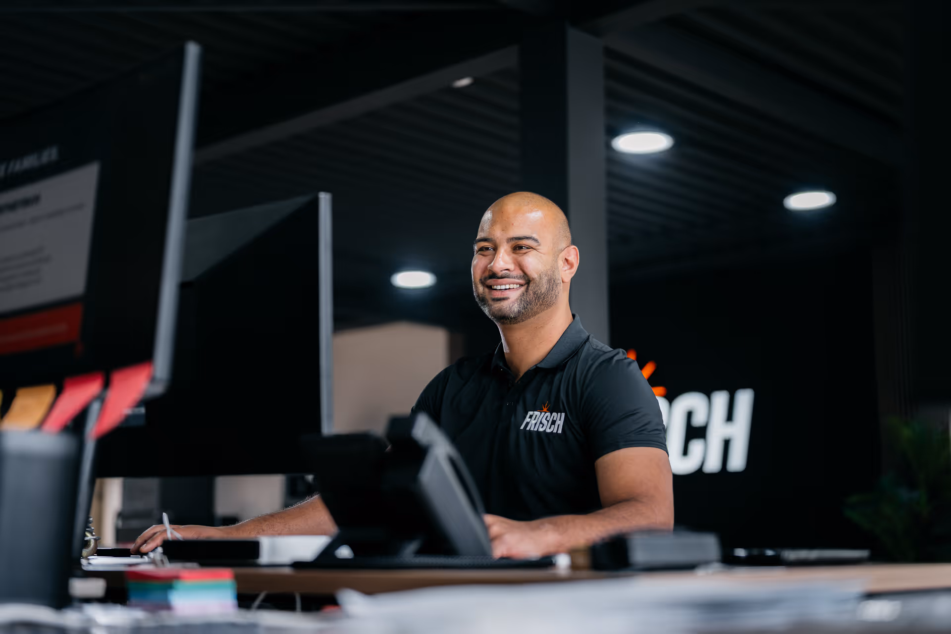 Lachende man achter bureau met computer in modern kantoor met Frisch-logo aan de muur.