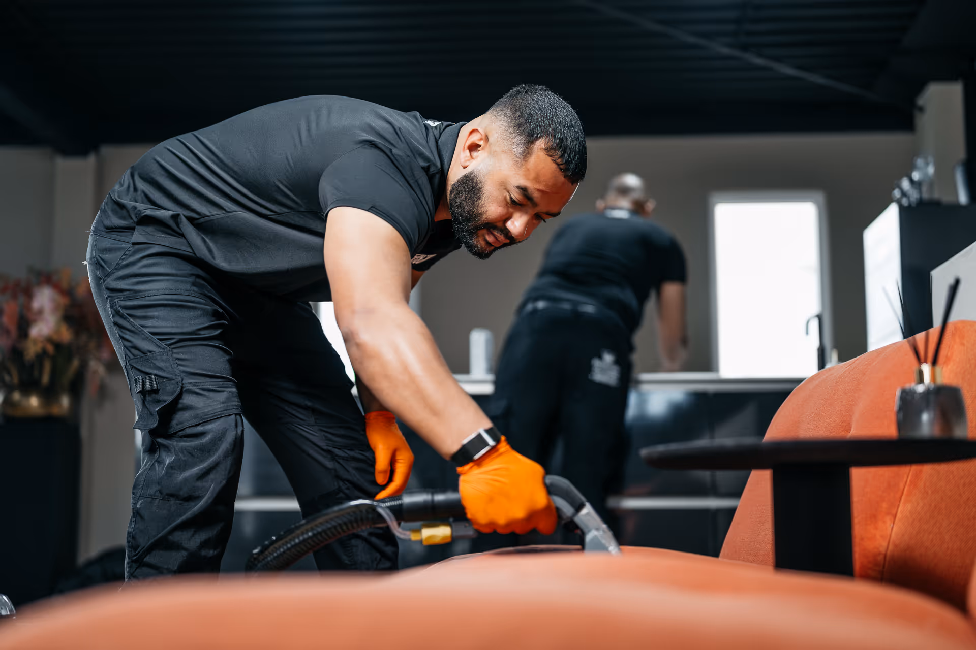 Man met oranje handschoenen reinigt een oranje bank met een stoomreiniger in een modern interieur.