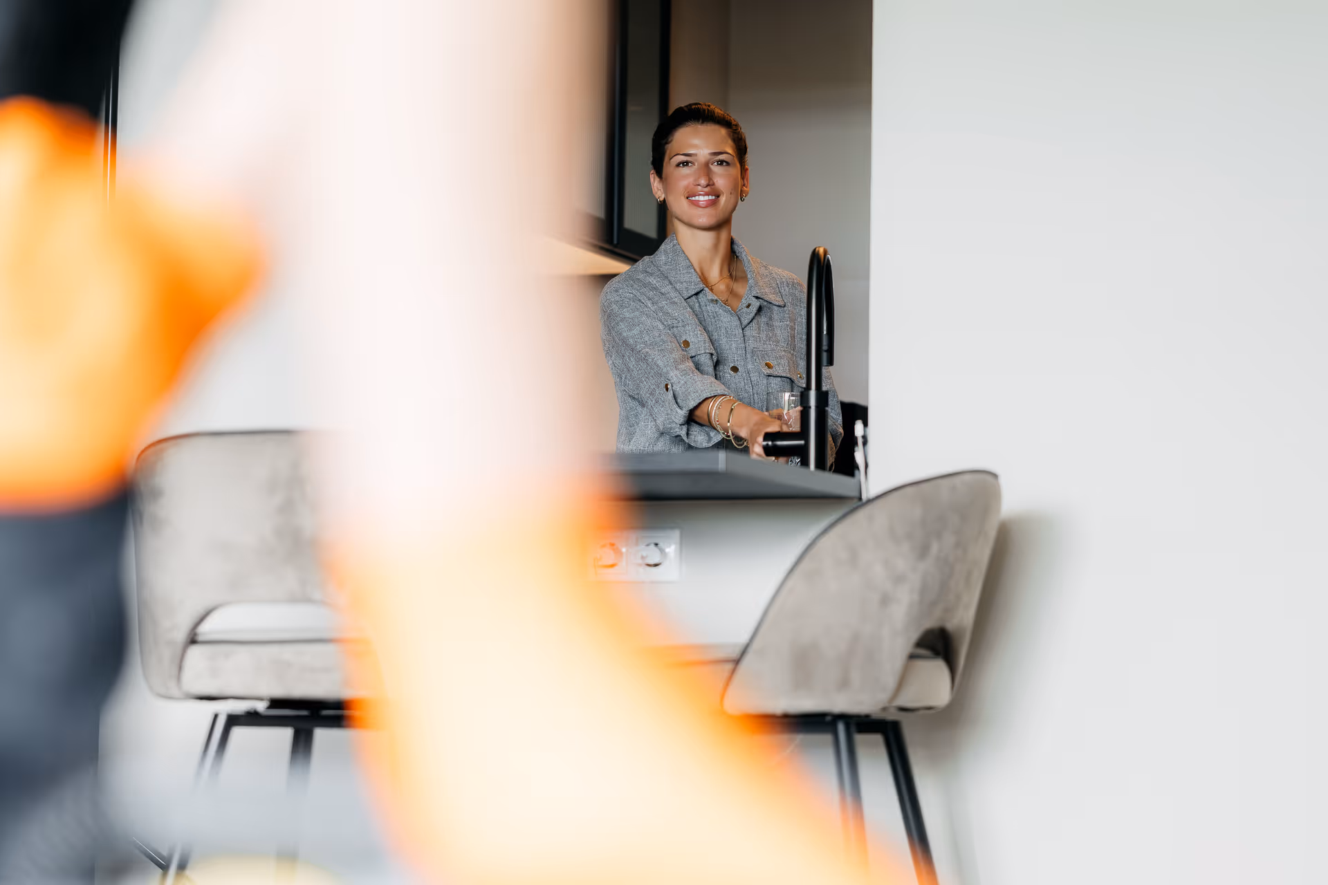 Vrouw in grijze blouse staat bij keuken aanrecht en glimlacht, wazig voorgrondobject in beeld.