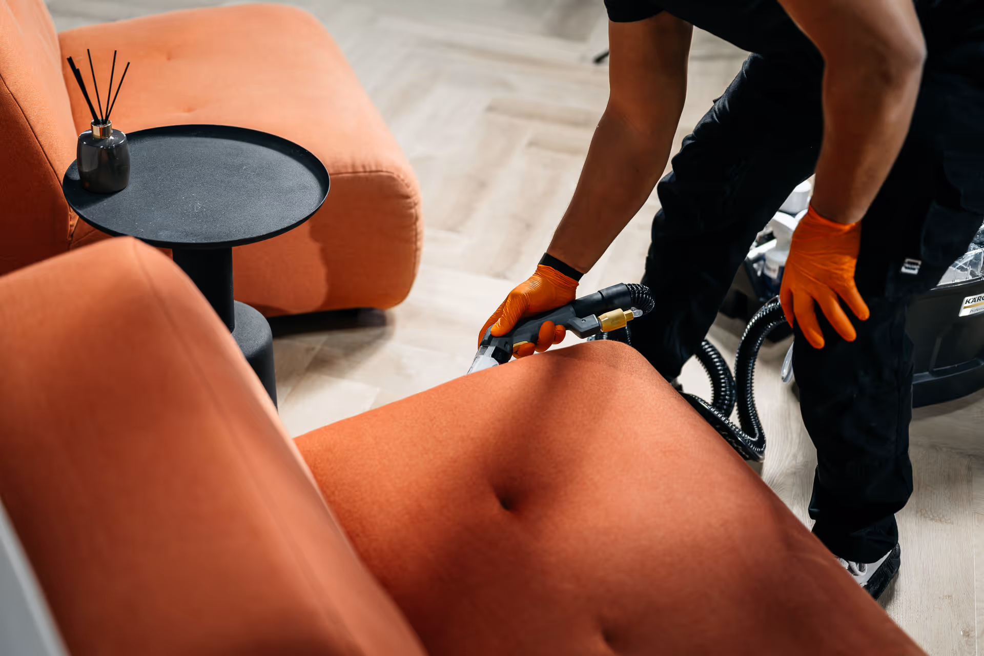 Persoon met oranje handschoenen die een oranje bankstof reinigt met een stoomreinigingsapparaat.