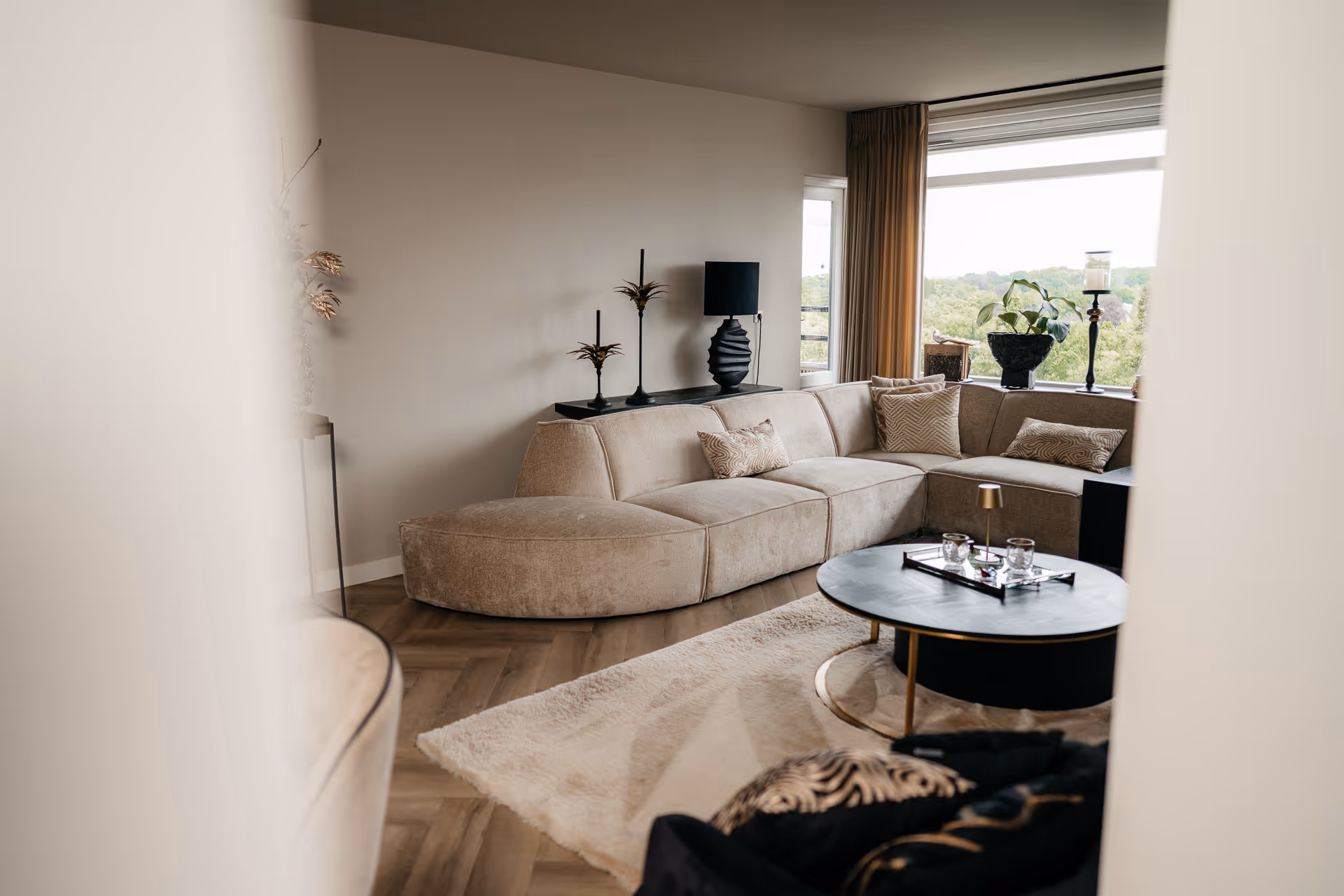 Moderne woonkamer met een beige hoekbank, zwart salontafel en een groot raam met uitzicht op groen.
