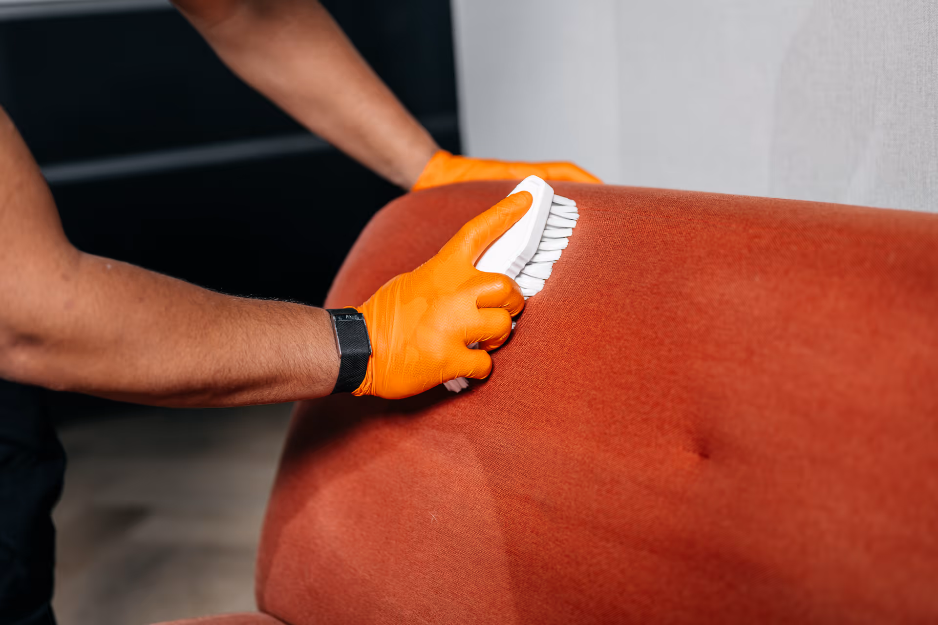 Iemand draagt oranje handschoenen en schrobt een rood gestoffeerde bank met een borstel.
